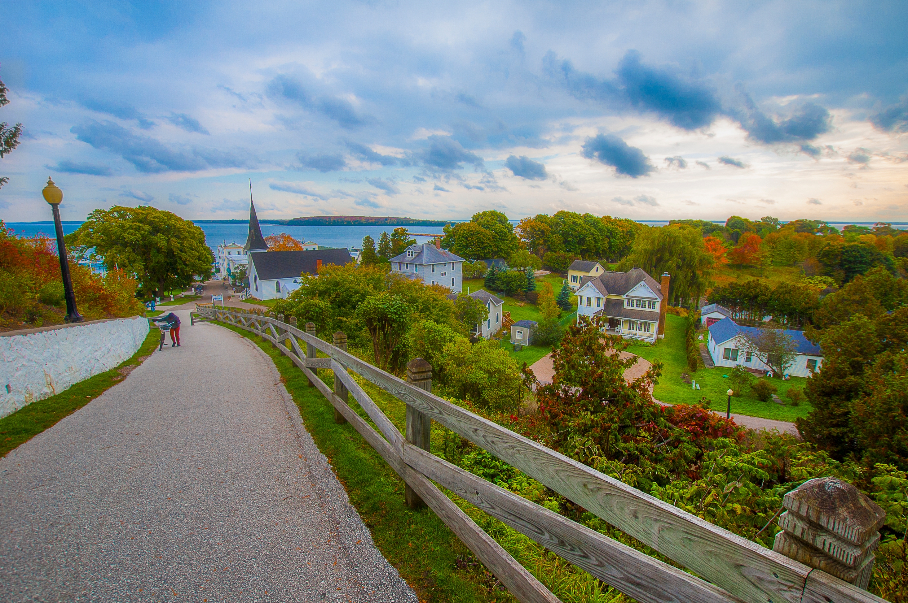 Scenic view of a quiet village with a church steeple, homes, and lush trees, along a wide path leading toward a distant body of water