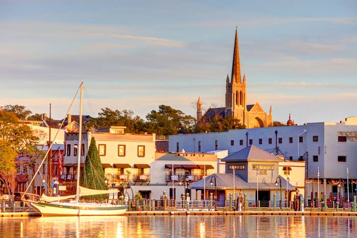 Harbor scene with sailboat docked by buildings and a church with a tall spire in the distance under evening light