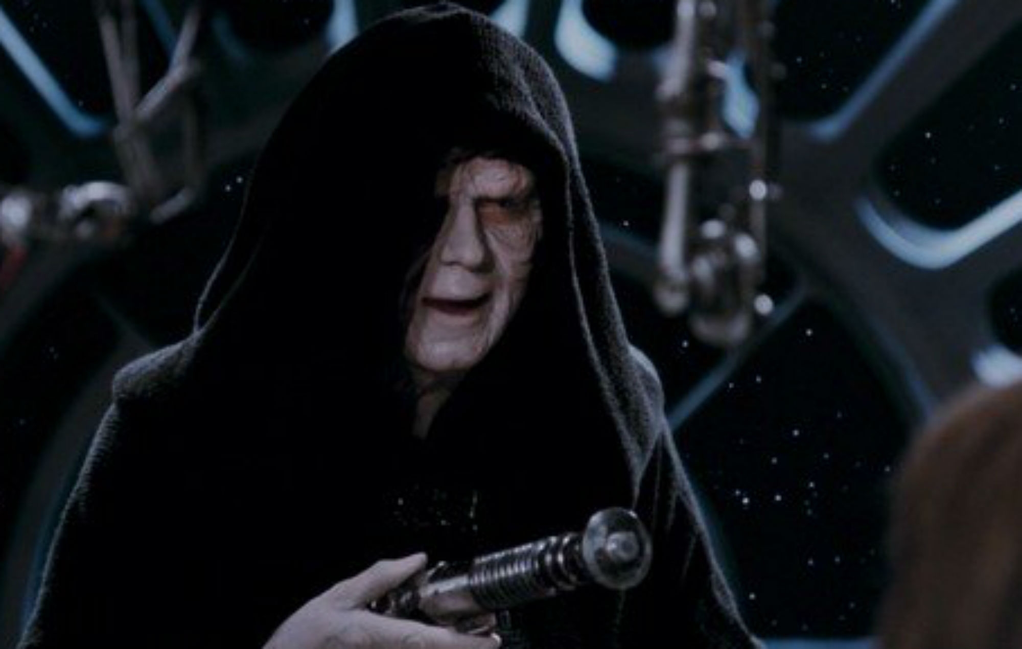 A character in a dark hooded robe holds a silver lightsaber hilt in a sci-fi setting with visible machinery