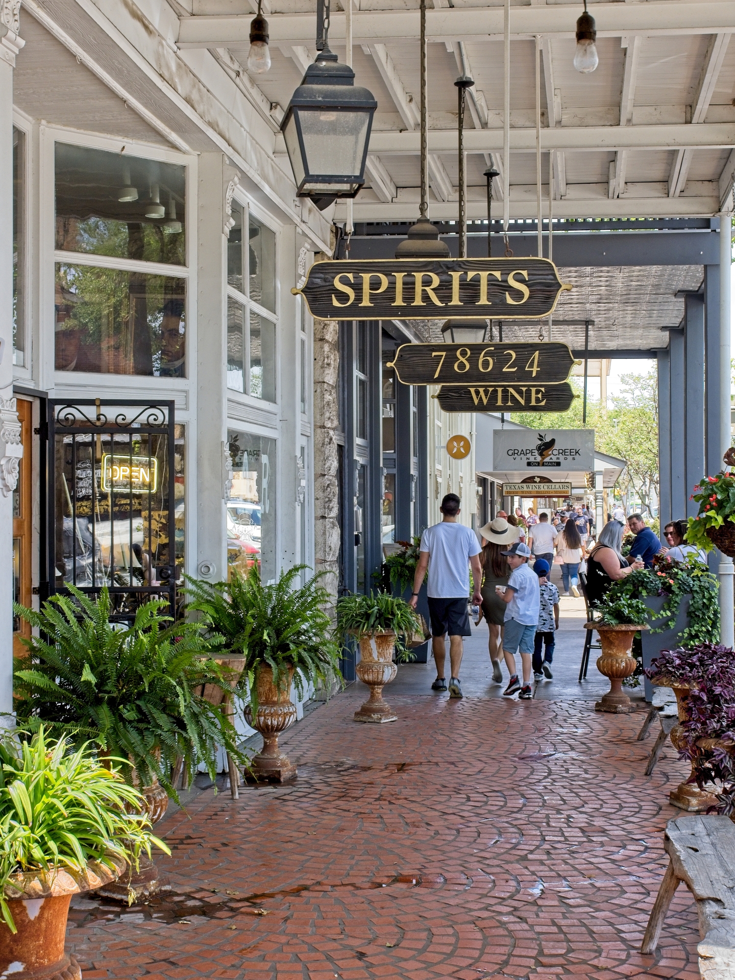 Pedestrians stroll along a bustling, plant-lined sidewalk beneath signs for spirits and wine