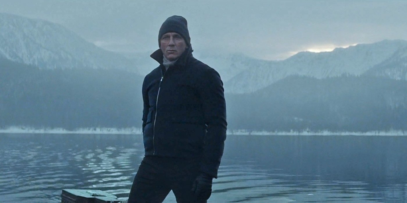 A person stands by a lake with snowy mountains in the background, wearing a beanie and a jacket in a cold, serene setting