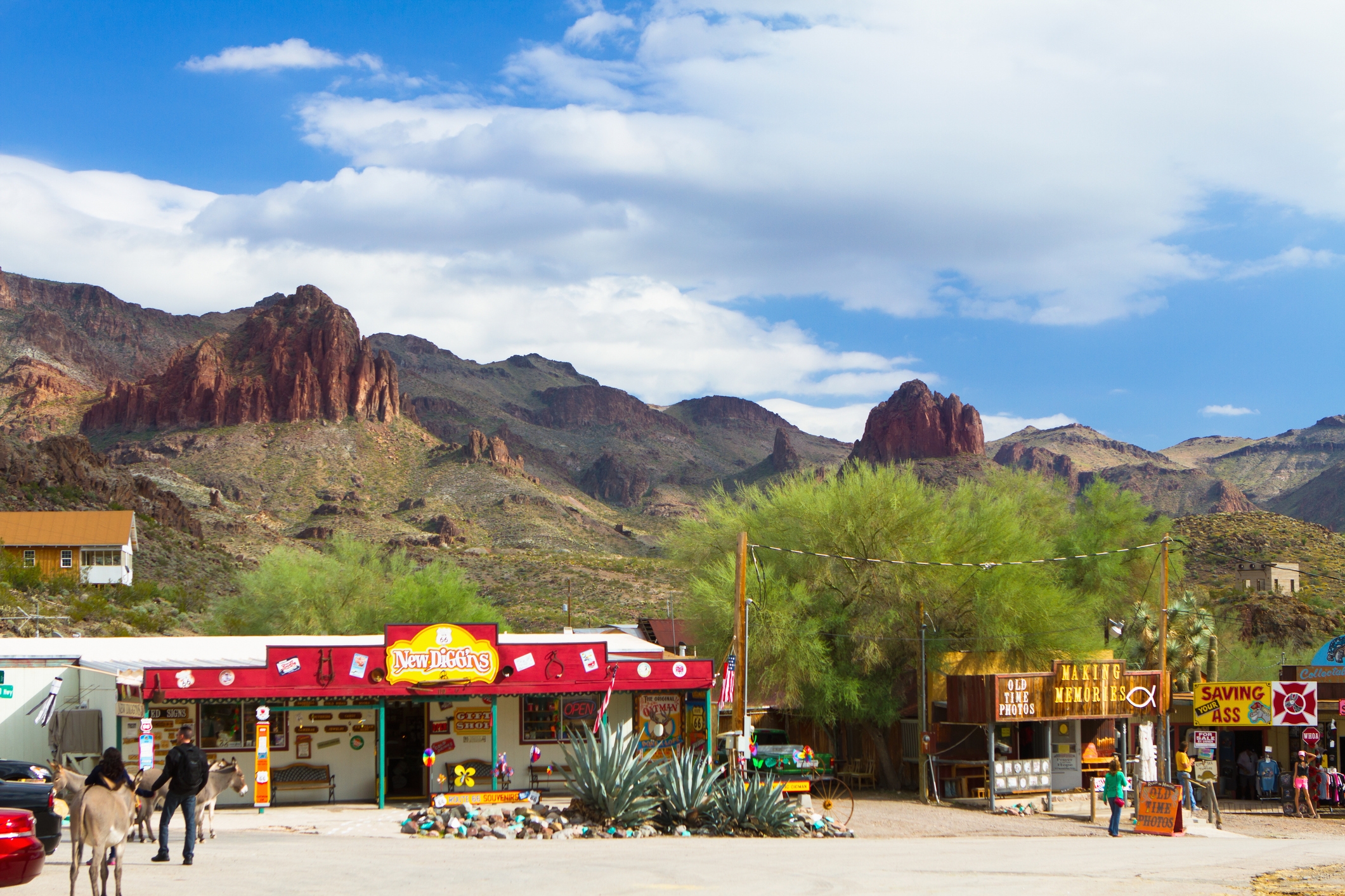Desert gas station with vintage signs, mountains in the background, a few scattered trees, and a person walking near a parked vehicle