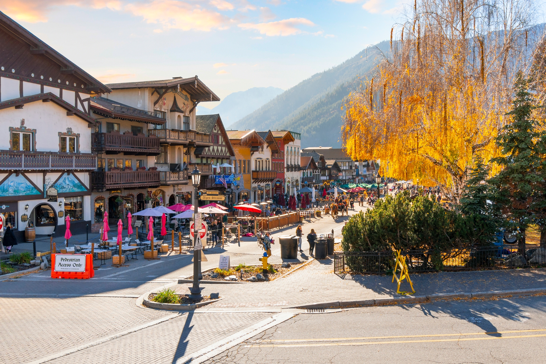 A busy, charming Bavarian-style village with shops, a restaurant patio, and people strolling along the street with mountains in the background