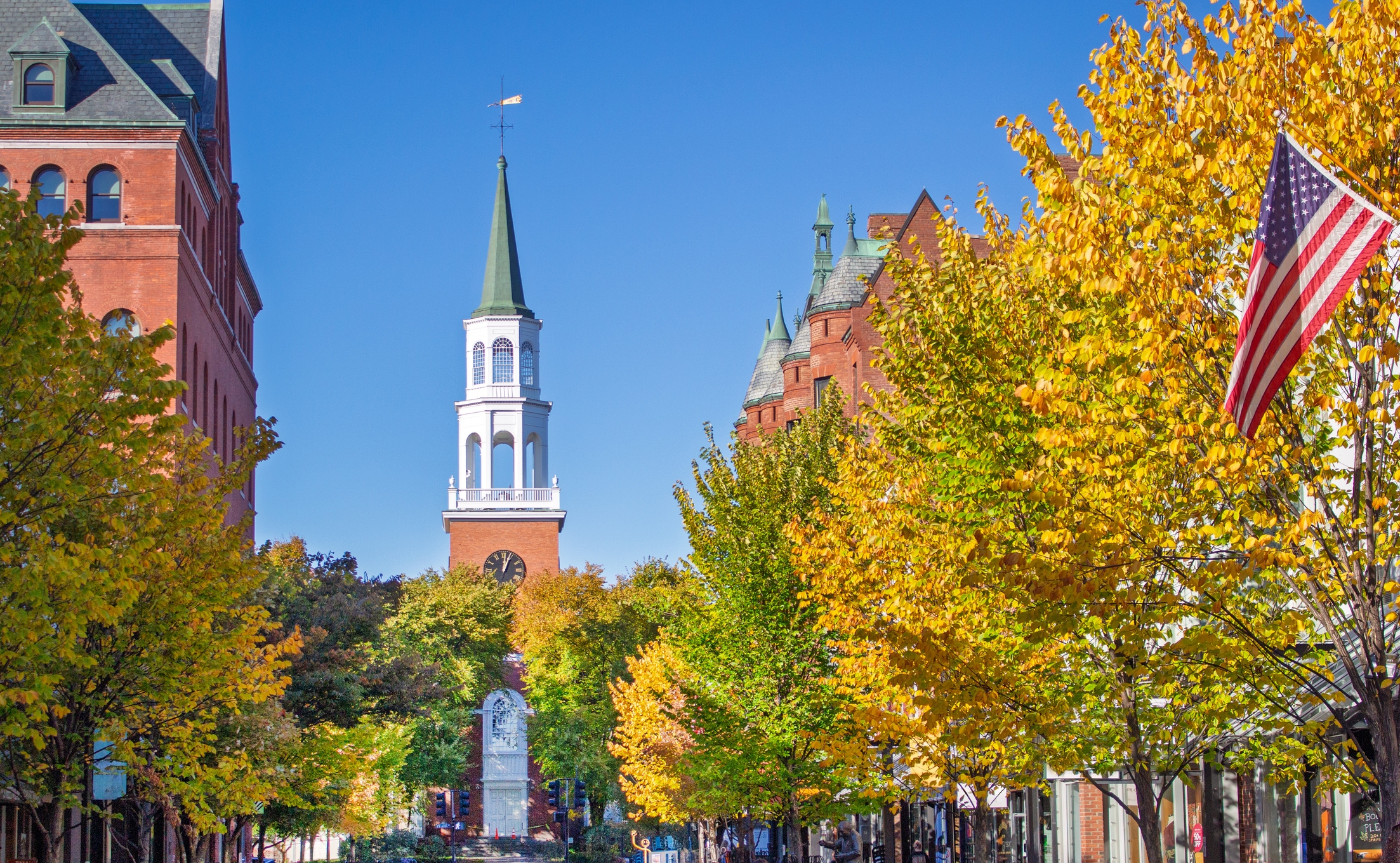 A scenic view of a street with colorful autumn trees, a church steeple in the background, and an American flag on the right