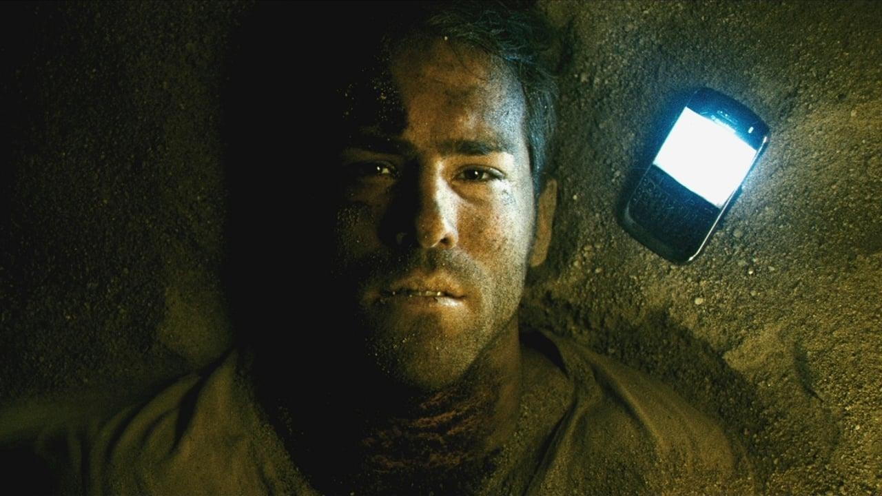 Man buried in sand with a dimly lit phone on his chest, appearing distressed. Scene suggests isolation or entrapment