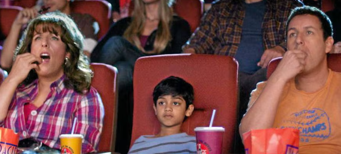 Three people sit in a movie theater, focused on the screen, with popcorn and drinks