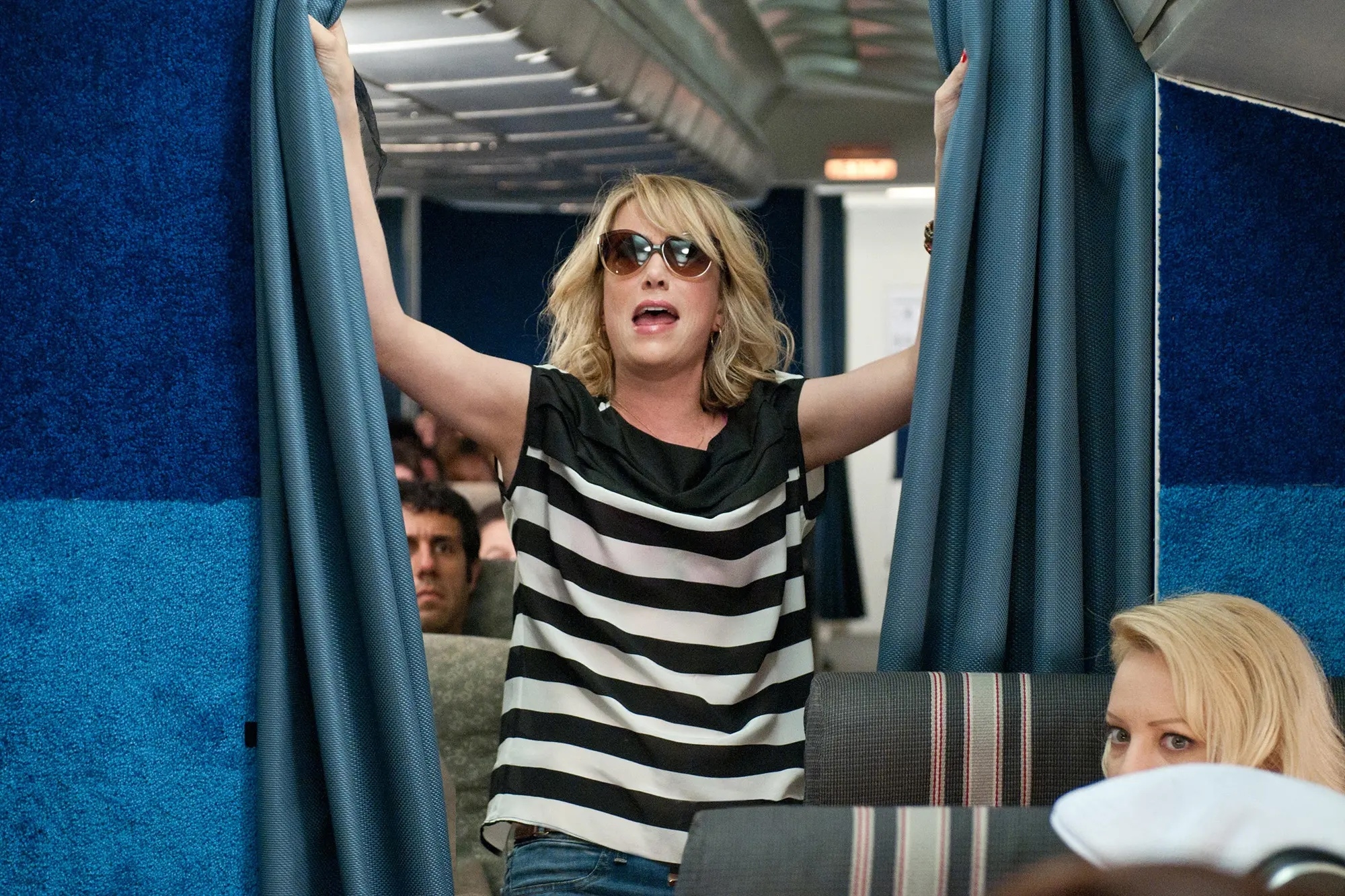 A person in sunglasses and a striped shirt dramatically holds open airplane curtains, with passengers looking on