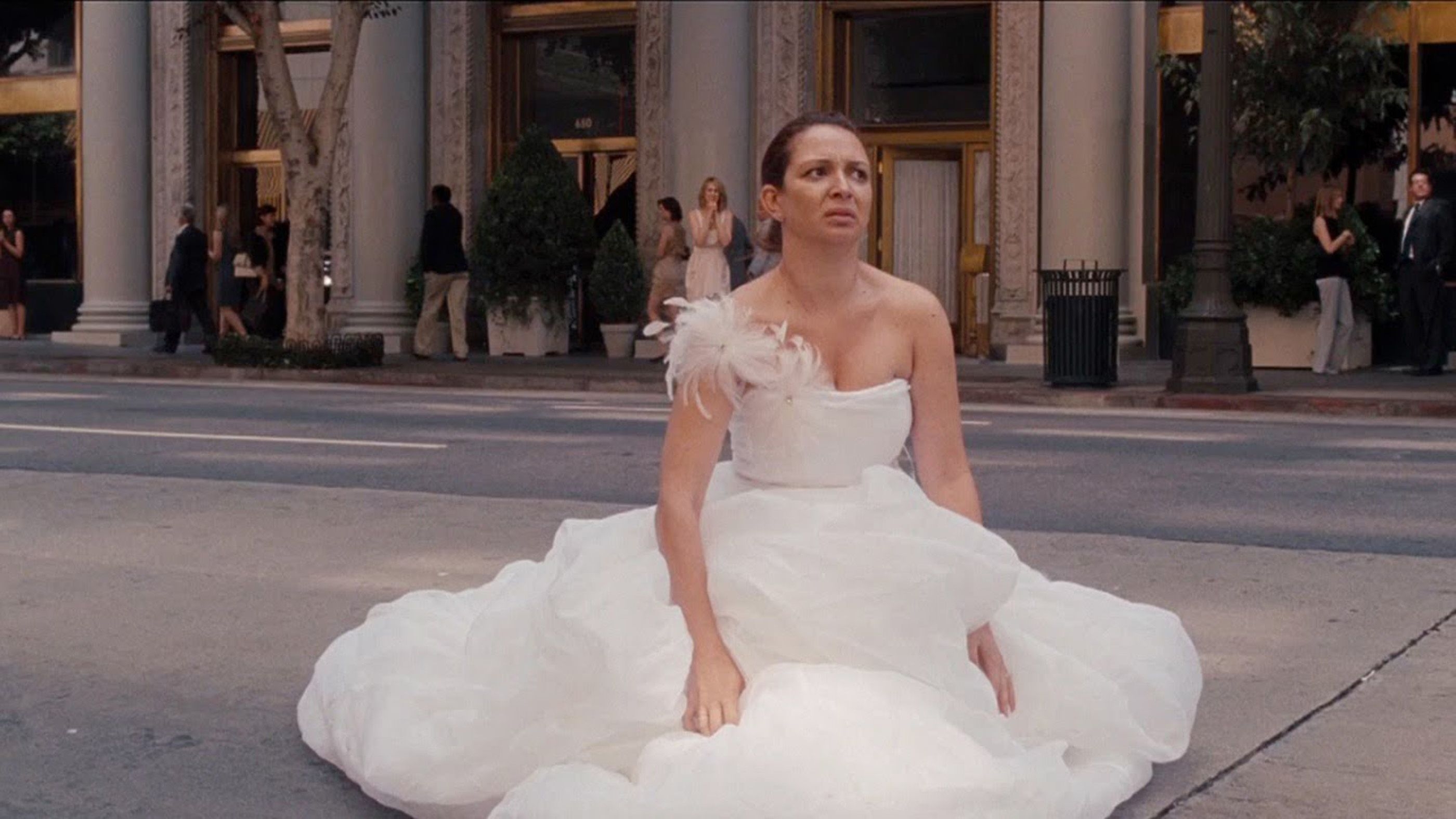 Person in a voluminous white gown sits on a city street, appearing contemplative, with people in formal wear in the background