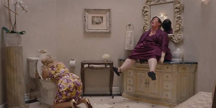 Two women in a bathroom scene; one kneels by a toilet, the other sits on a sink, both possibly in distress