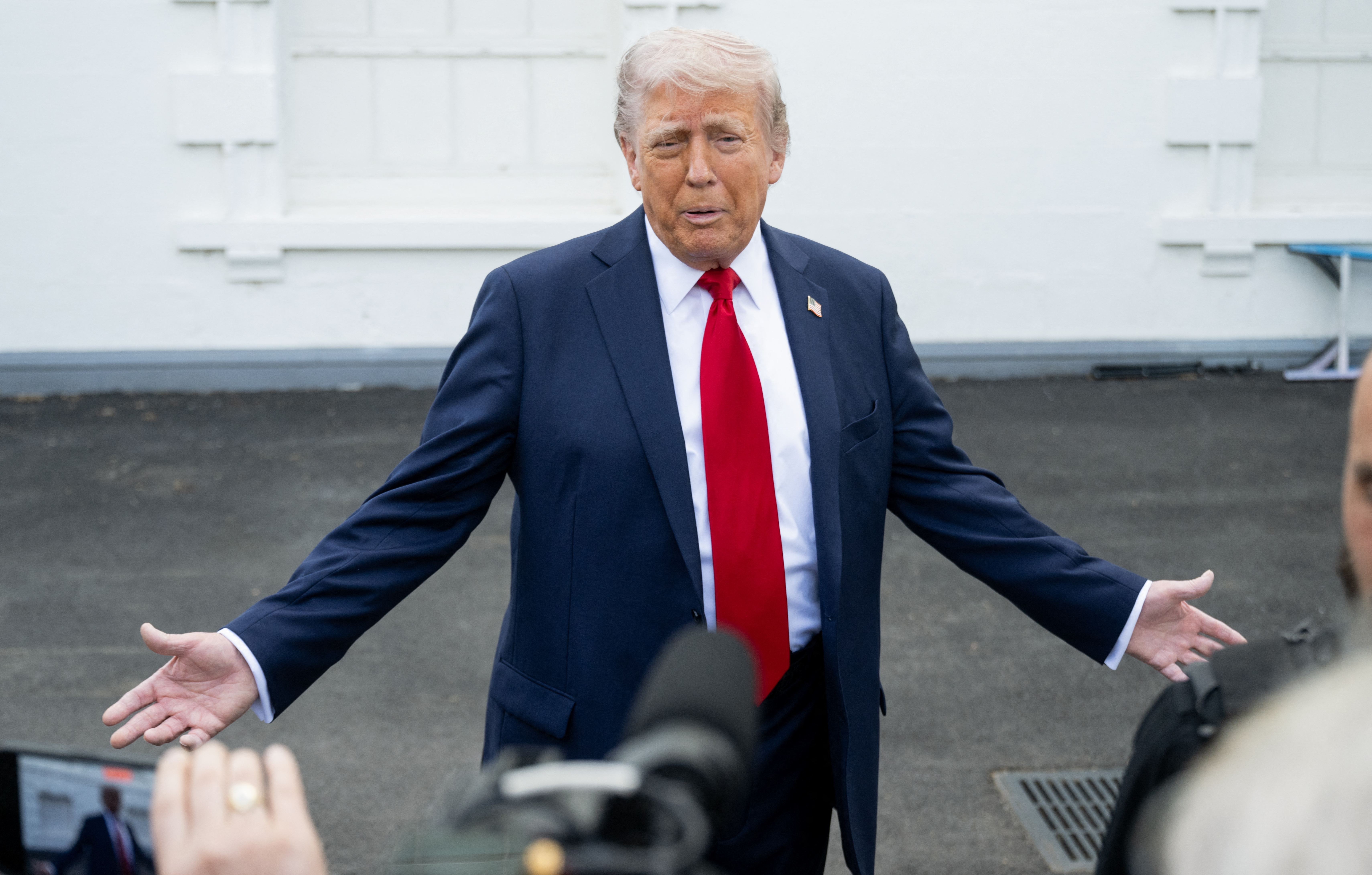 A person in a suit and red tie gestures with open arms, speaking to reporters outside a building. Microphones are visible in the foreground