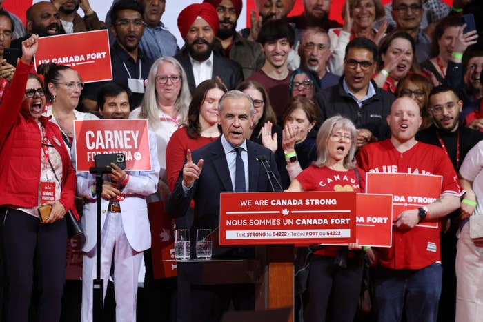 A person speaks at a Canada Strong event, surrounded by supporters holding signs and wearing casual attire