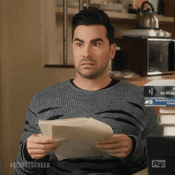 A surprised man in a casual sweater holds papers, looking up with wide eyes and raised eyebrows