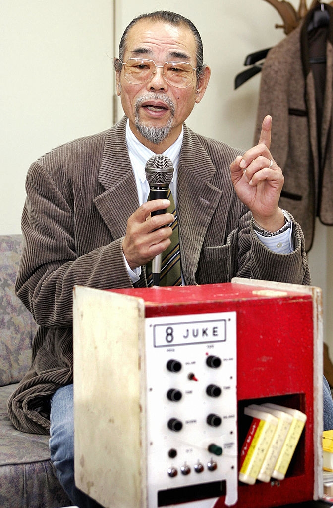 Inoue speaks into a microphone while sitting next to a vintage karaoke machine labeled "8 Juke"