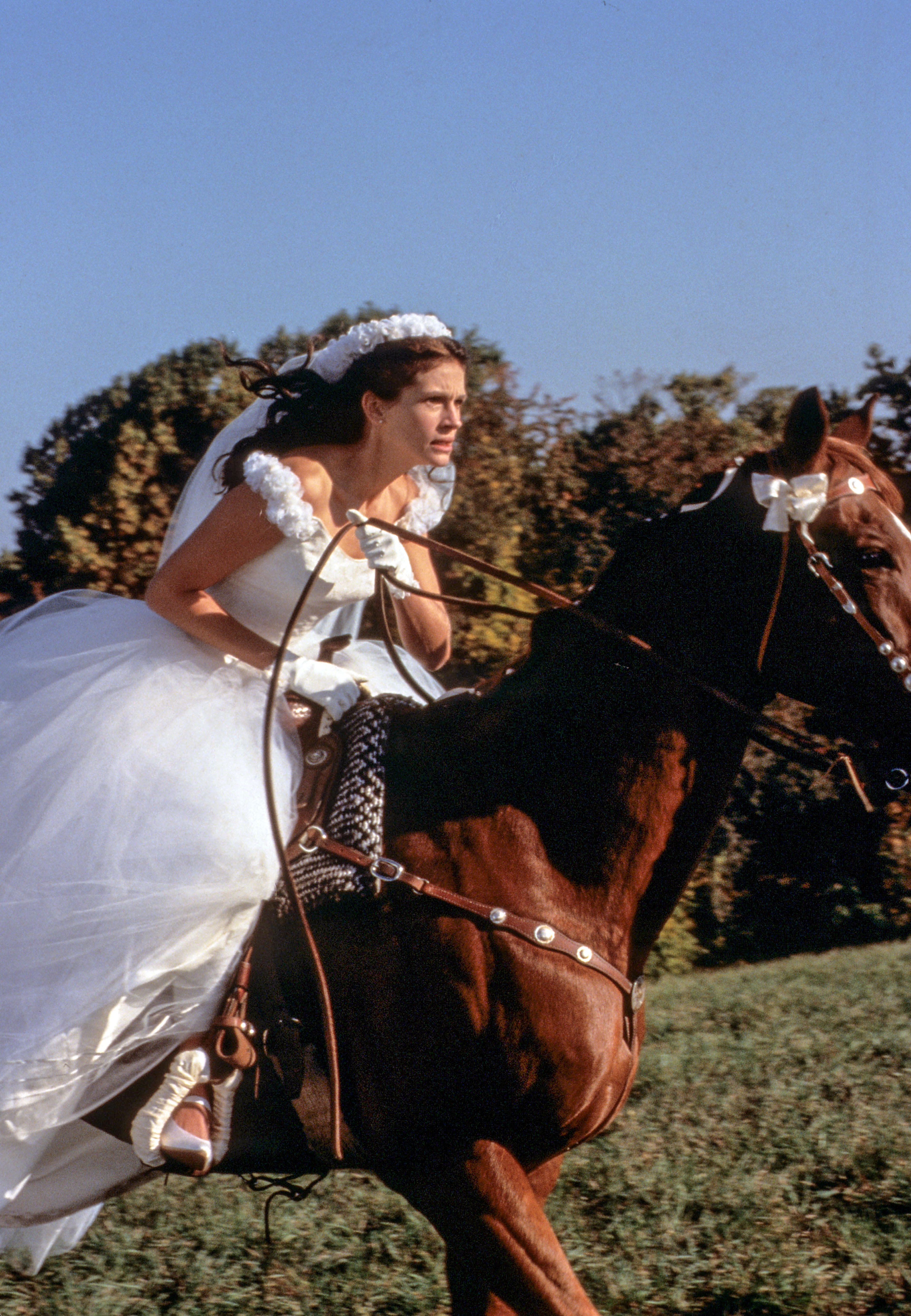 A woman in a wedding dress rides a horse outdoors, looking determined