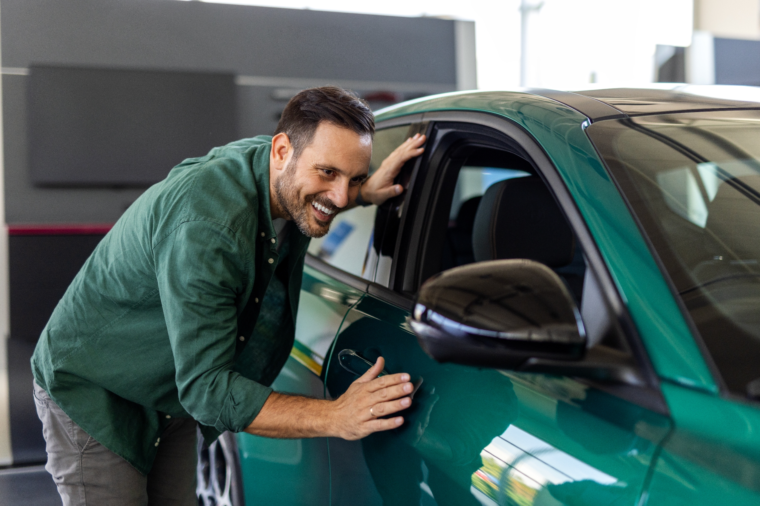 A smiling man in a casual long-sleeve shirt inspects a car at a showroom, touching the door lightly