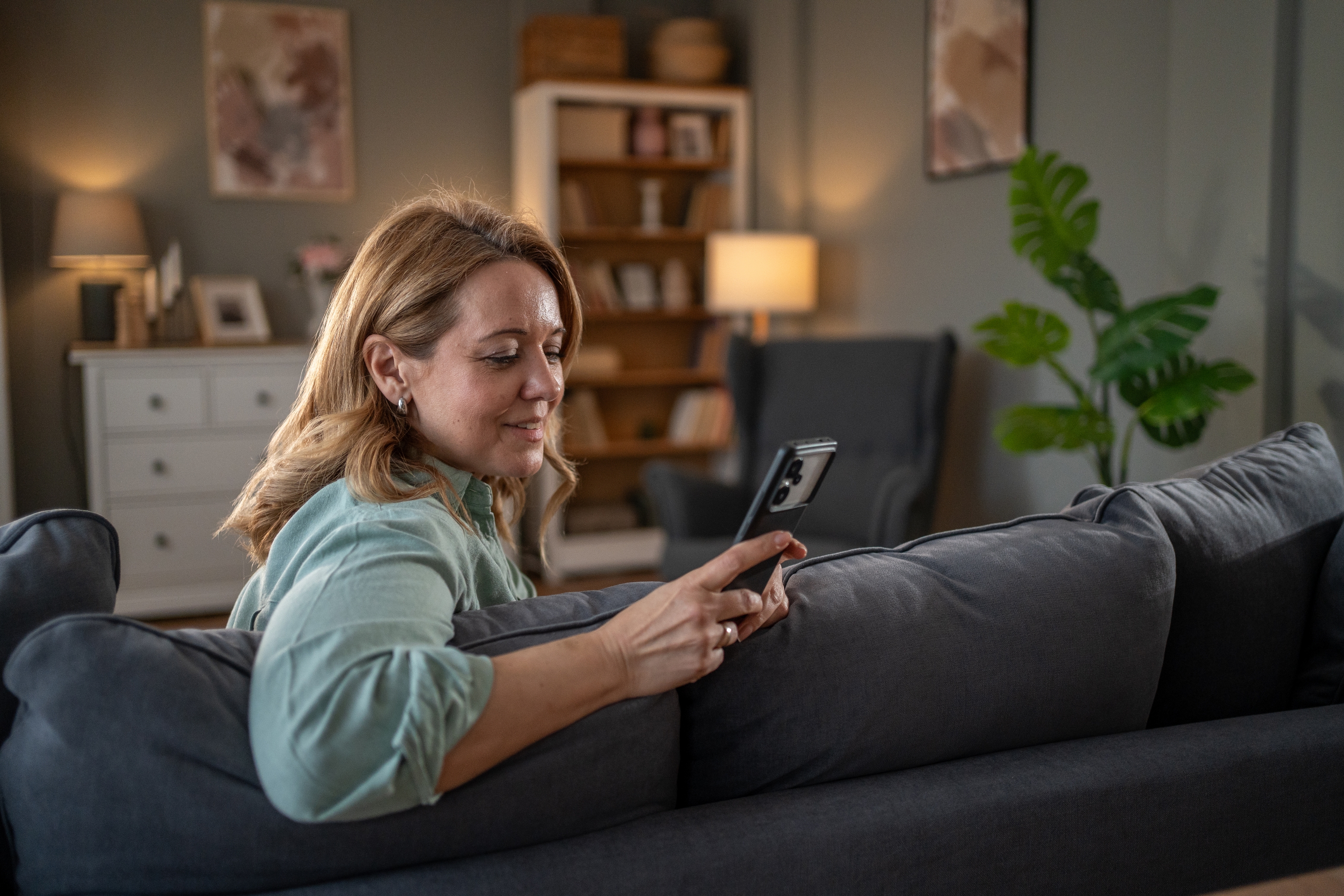 A person sitting on a couch, looking at a smartphone, with a cozy living room scene in the background