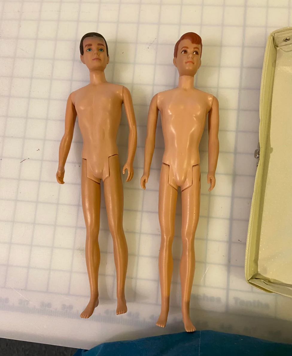 Two vintage male dolls, unclothed, side by side on a grid surface. The one on the left has darker, more cropped hair, while the one on the right has lighter reddish hair and a sort of side bang