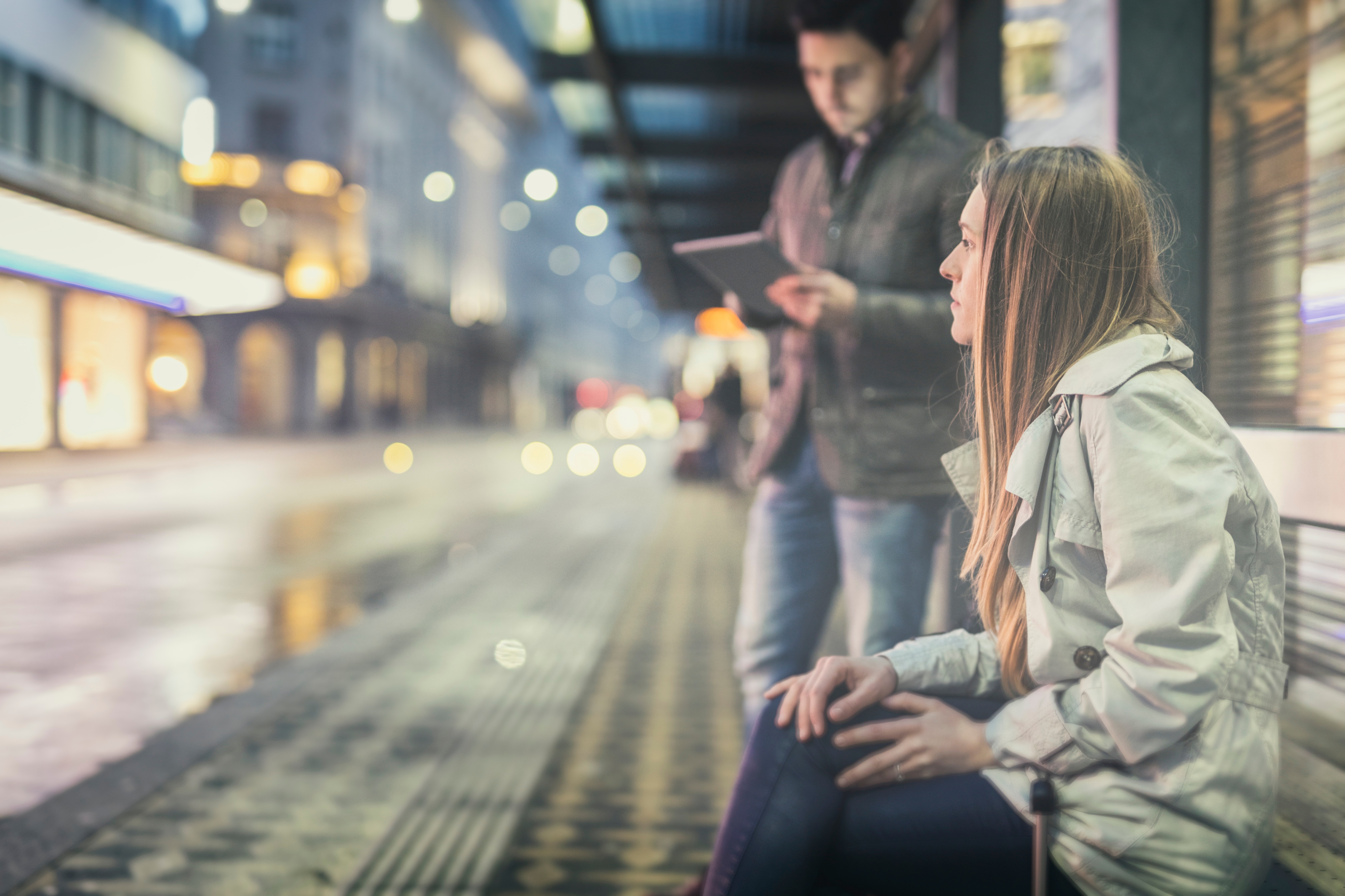 Woman sitting at a bus stop bench looking thoughtful, while a man stands nearby using a tablet. City lights and traffic are blurred in the background