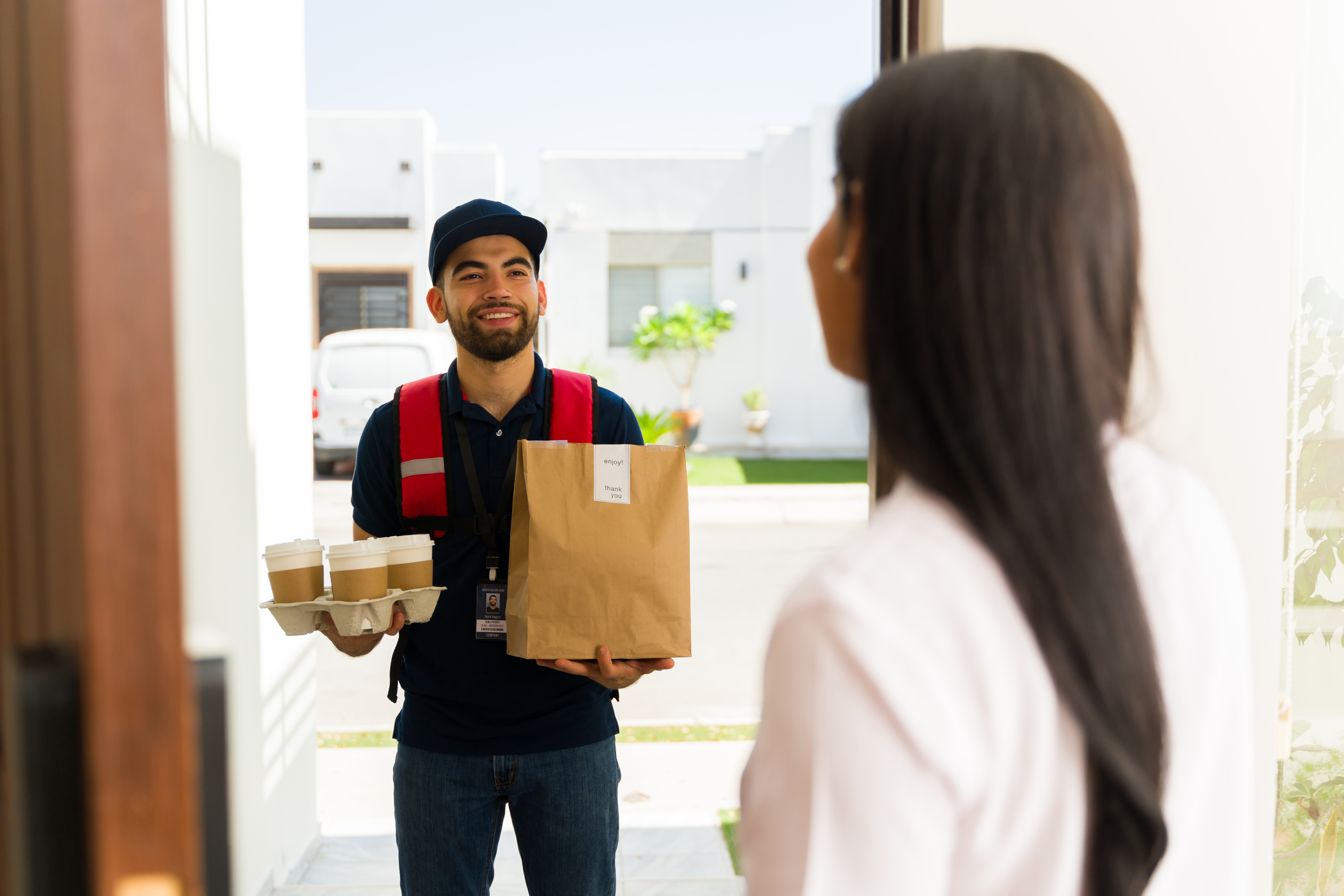 A delivery person smiles while handing a customer coffee cups and a paper bag at the door