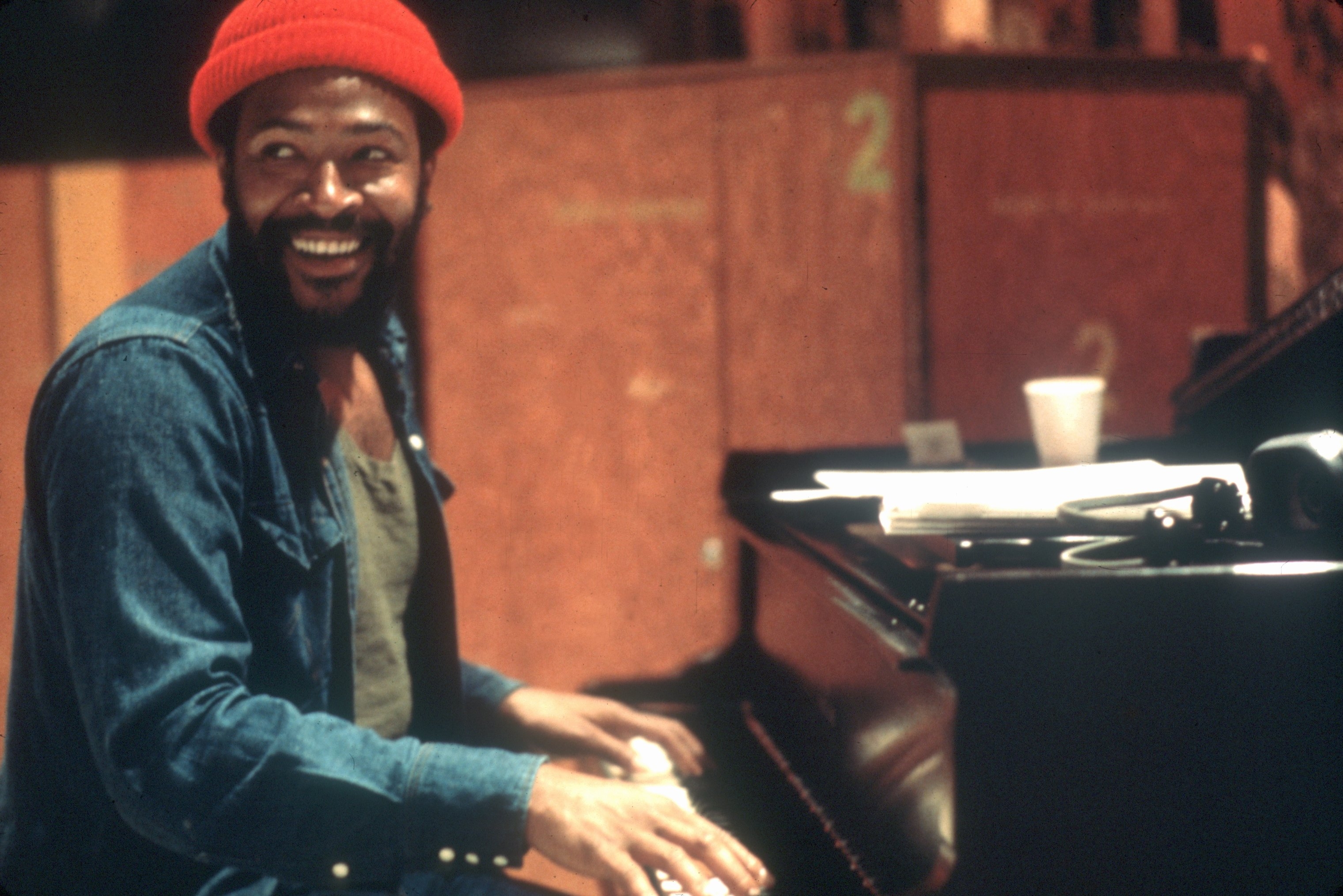 Marvin Gaye wearing a denim jacket and red beanie smiles while playing the piano