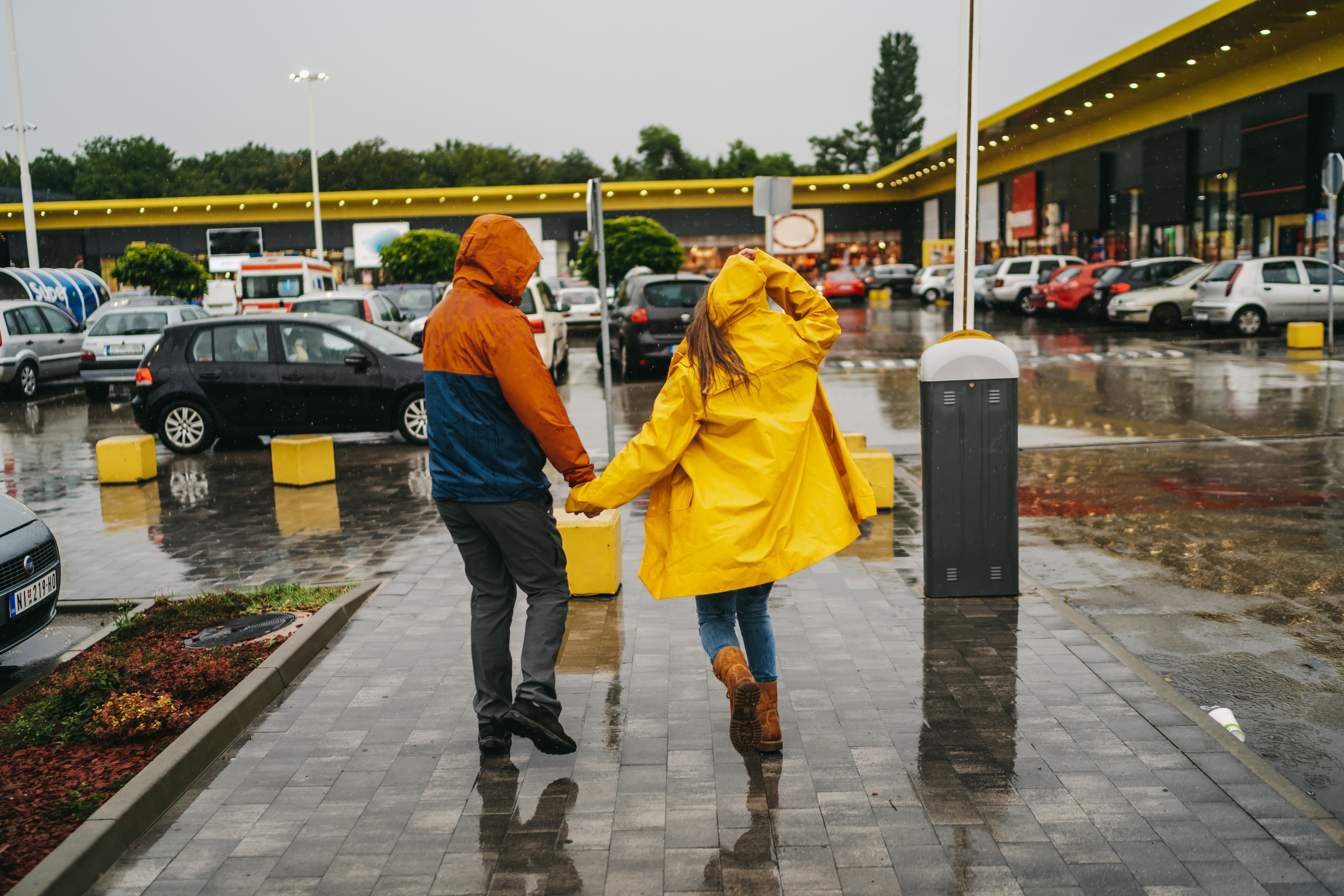 Two people in rain jackets walk hand-in-hand through a wet parking lot, shielding themselves from the rain