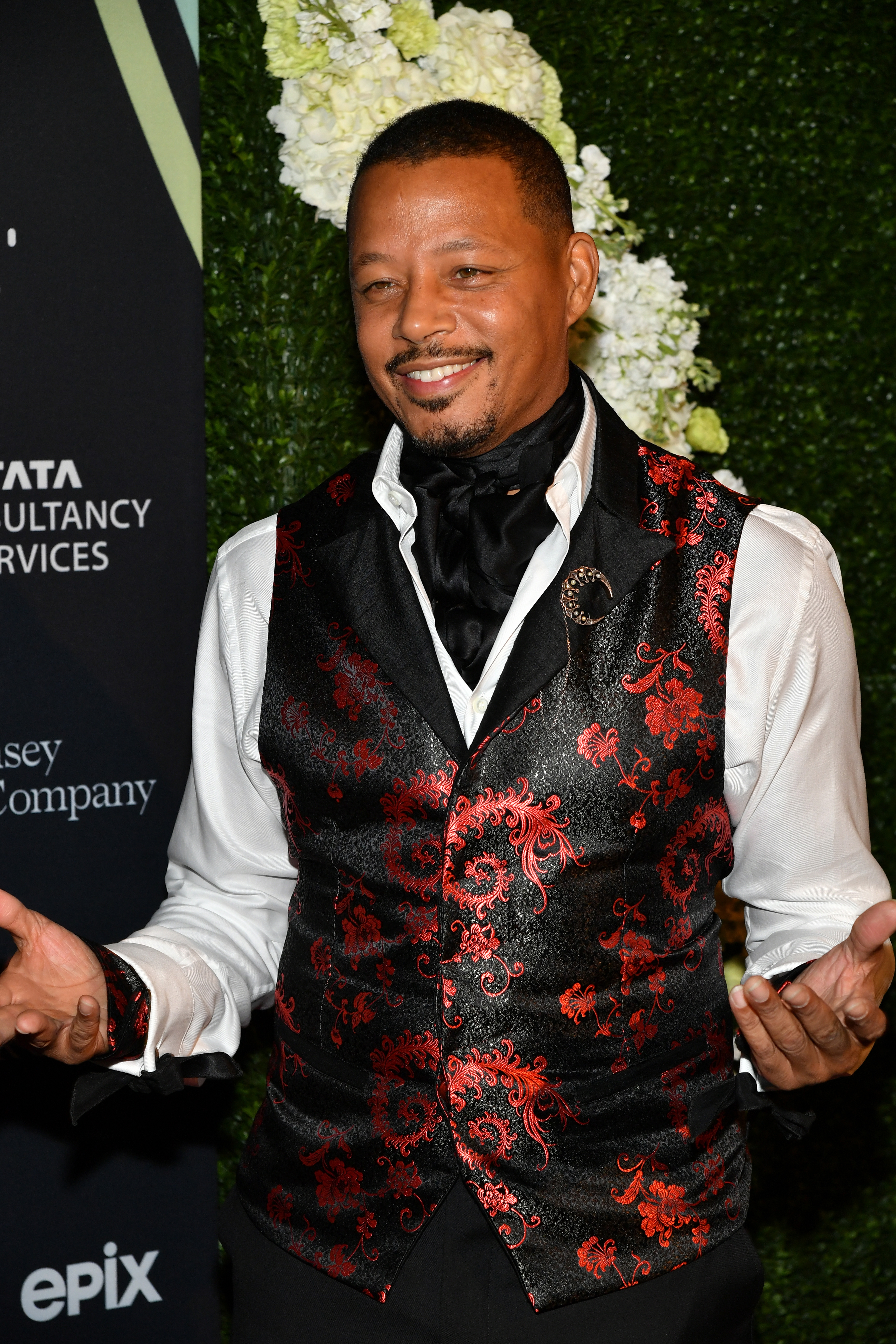 Terrence Howard in an elegant red and black patterned vest with a white shirt and black cravat, standing against a floral backdrop