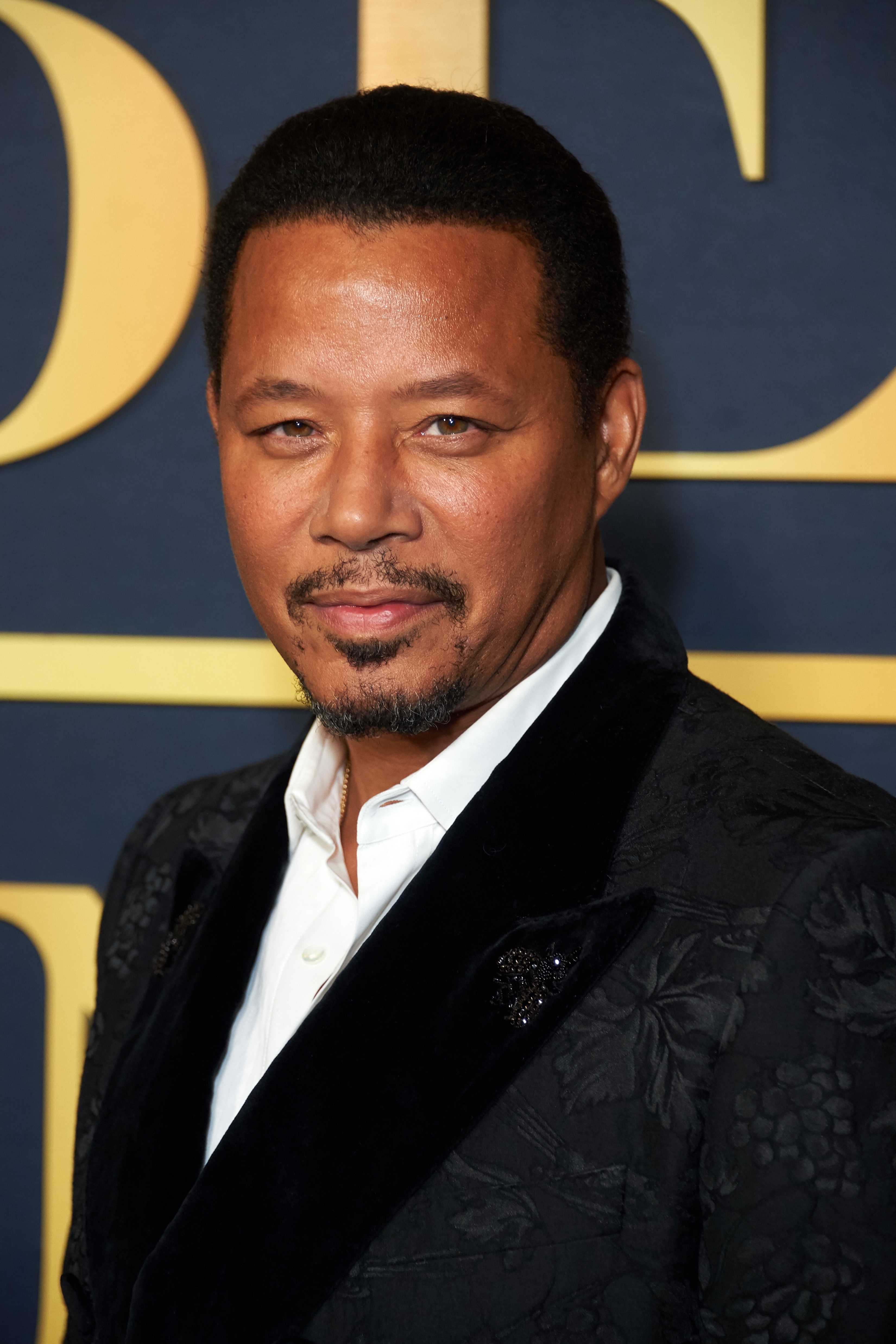 Terrence Howard at a formal event wearing a dark patterned jacket and white shirt, posing in front of a branded backdrop