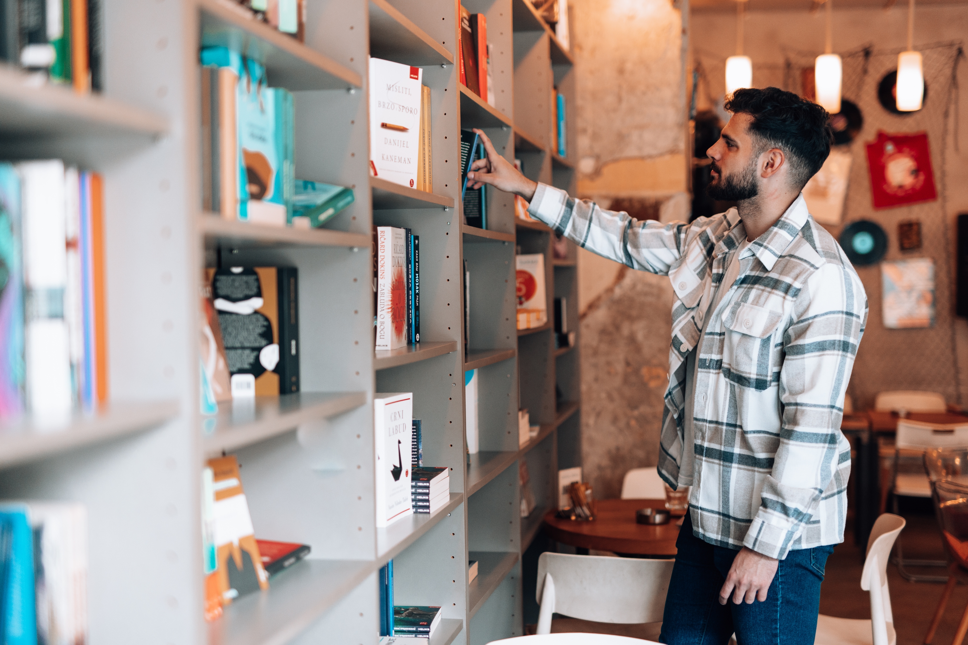 Person in a casual plaid shirt browsing books on a bookstore shelf. Cozy interior with seating area in the background