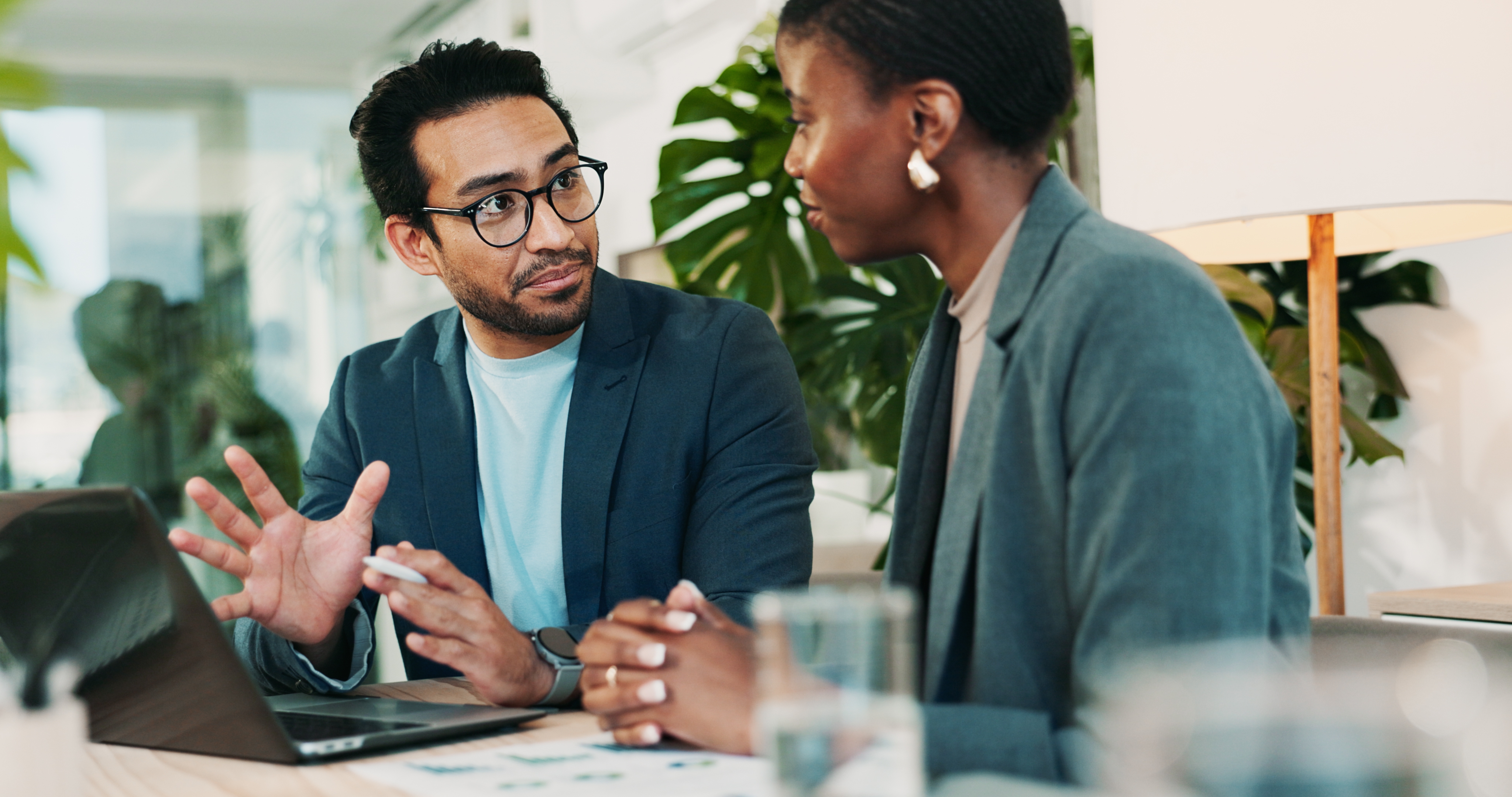 Two people in business attire have a discussion at a desk, one gesturing toward a laptop, suggesting collaboration or brainstorming