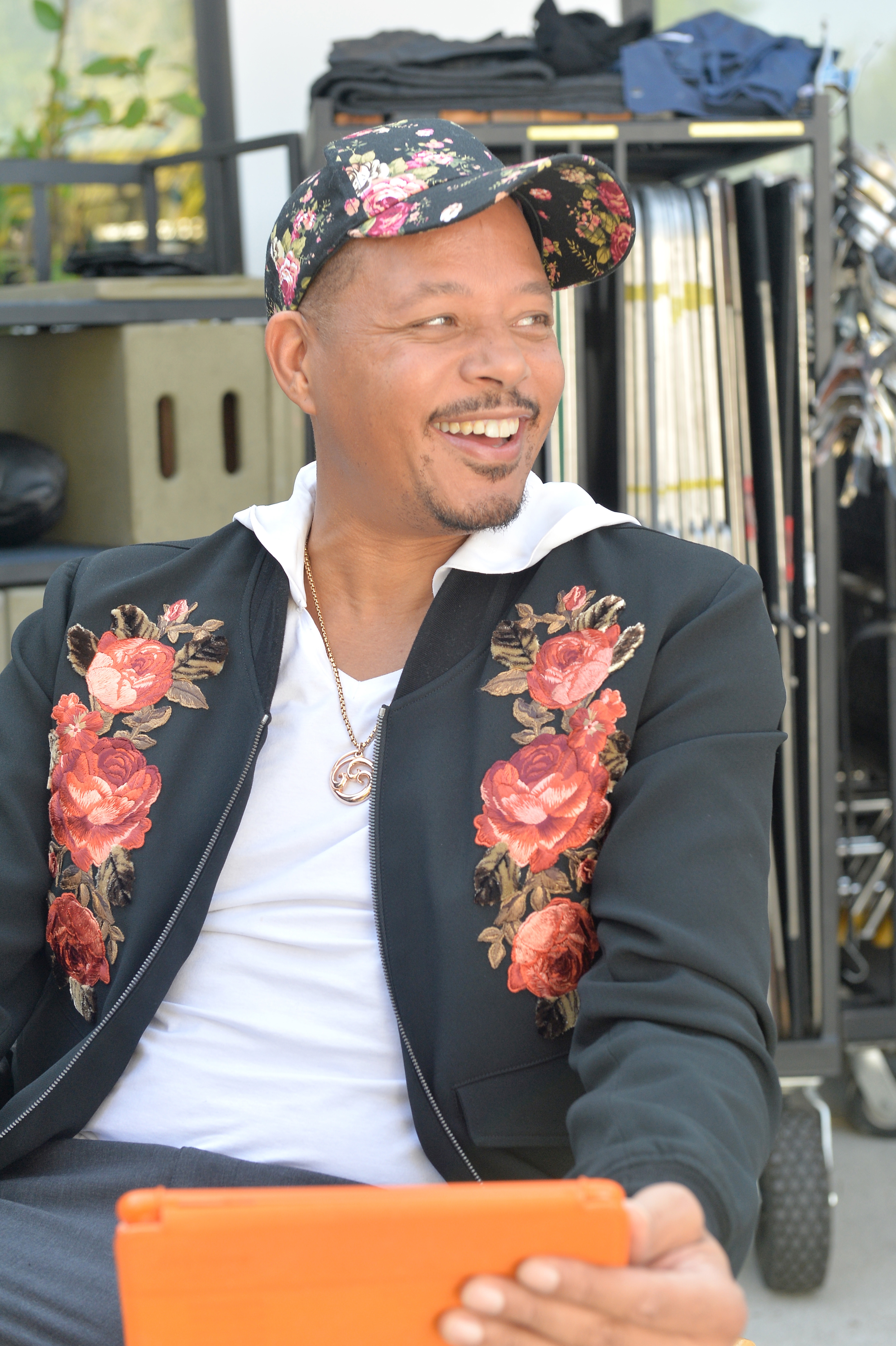 Terrence Howard sits and smiles, wearing a floral hat and jacket with rose patterns, holding a tablet