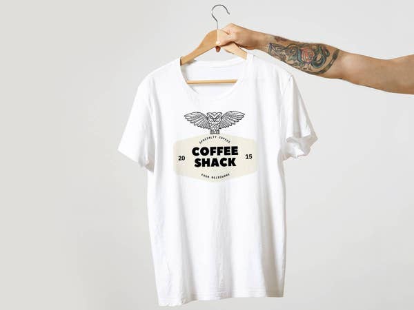 White T-shirt on hanger with \"Coffee Shack\" graphic featuring an owl and the year 2015. Arm with tattoo holds the hanger