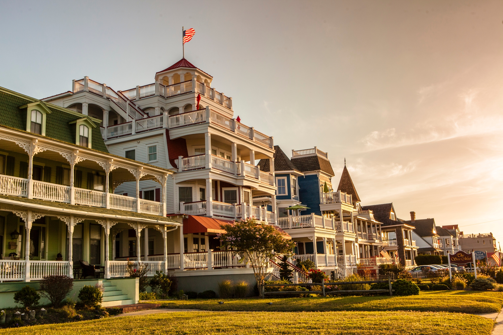 Victorian-style homes with intricate balconies line a street, with a sunset in the background and a small American flag atop one building