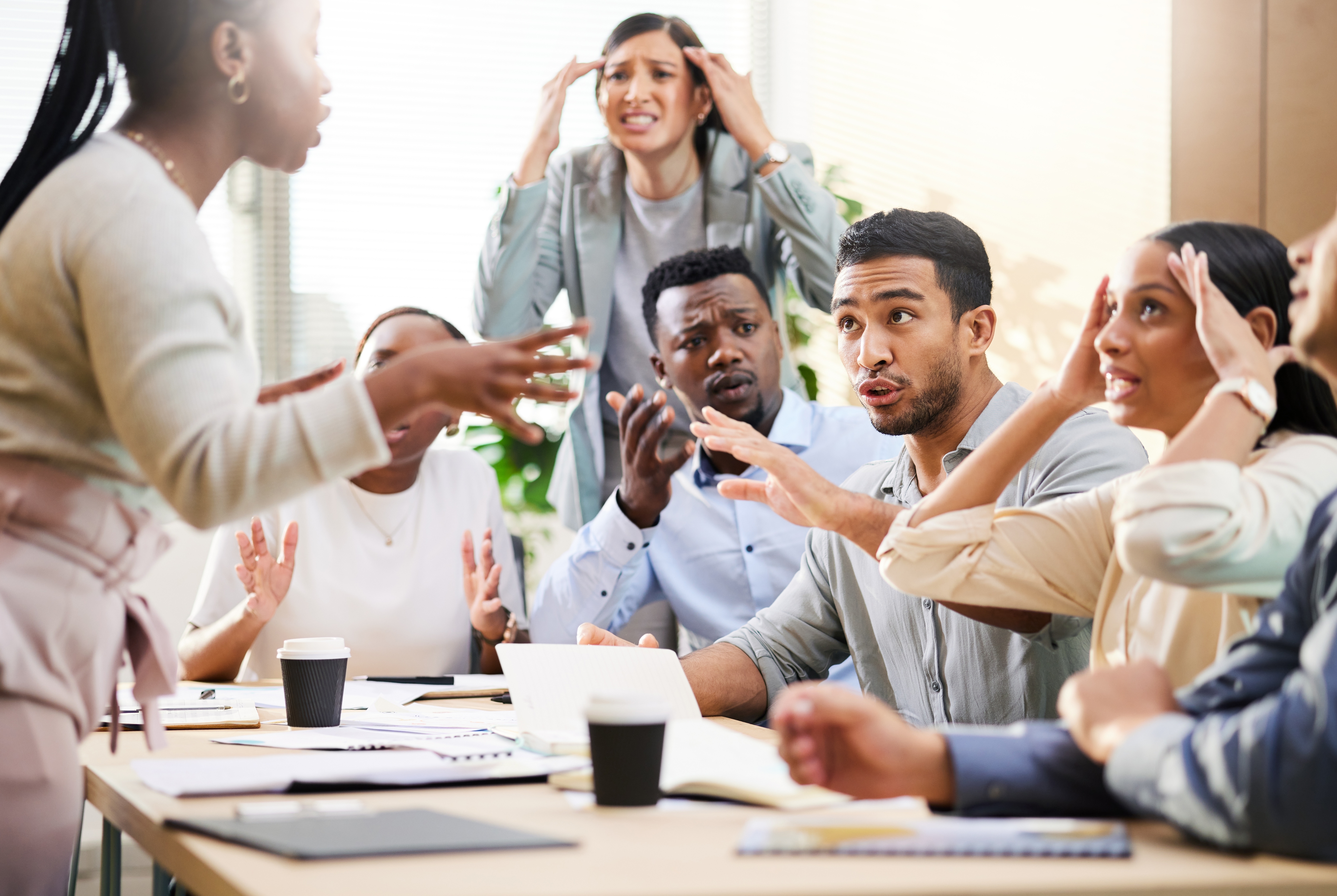 People in an office meeting appear frustrated and animated, gesturing and discussing intensely around a table with coffee cups and papers