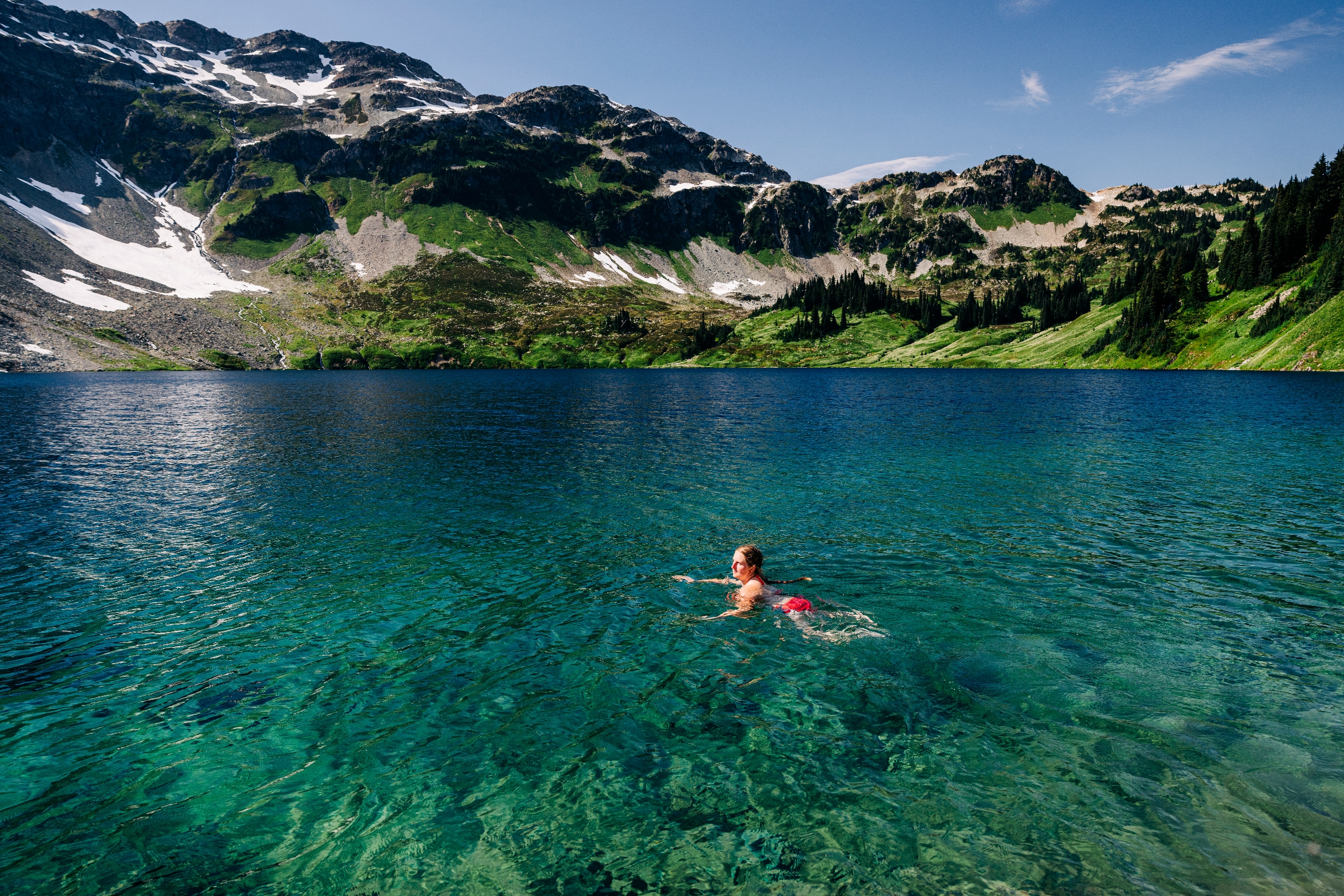 Person swimming in a clear mountain lake with distant snowy peaks and lush greenery in the background