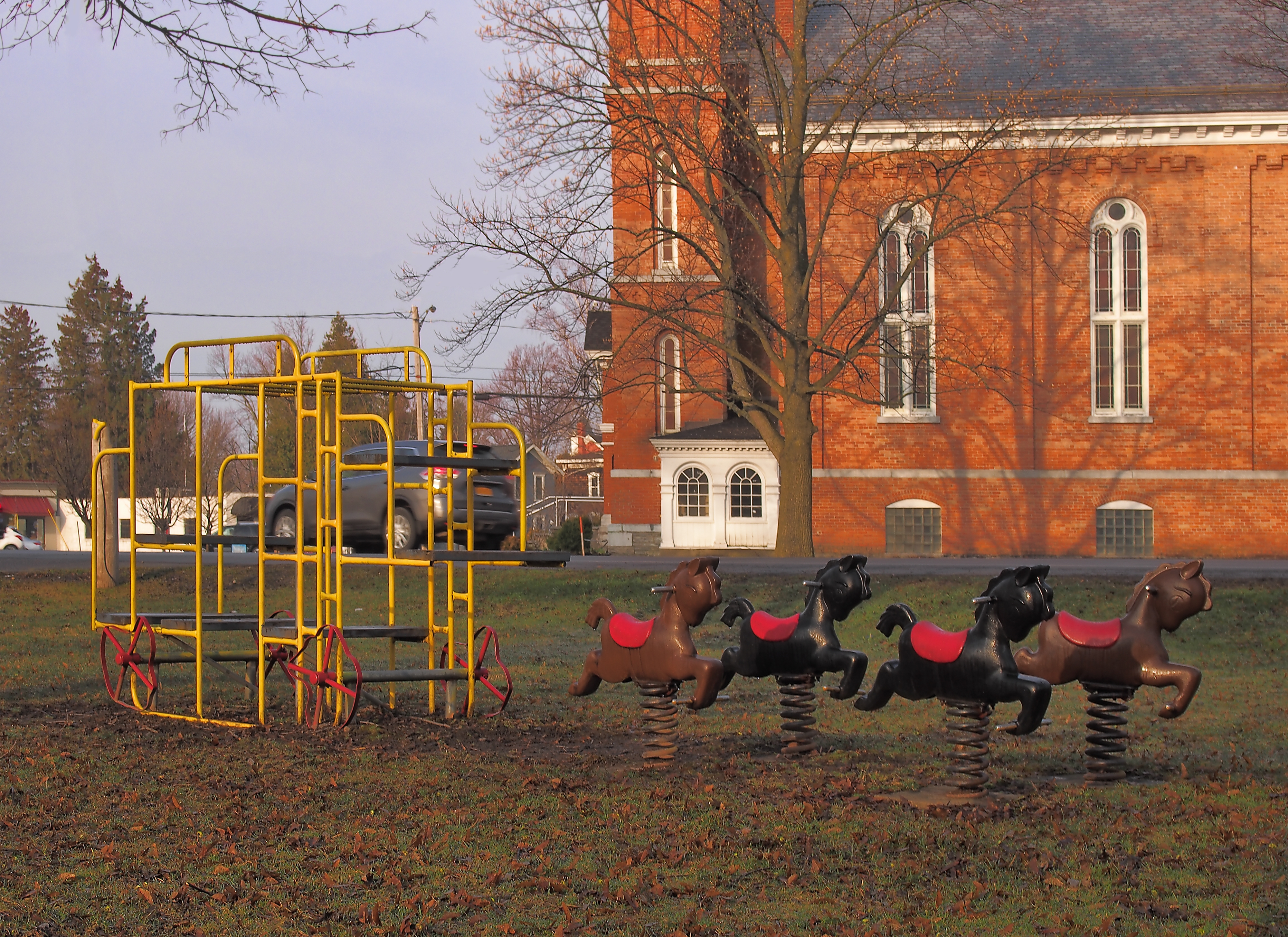 Playground with a vintage metal slide and horse spring riders in front of a historic brick church with arched windows and a leafless tree
