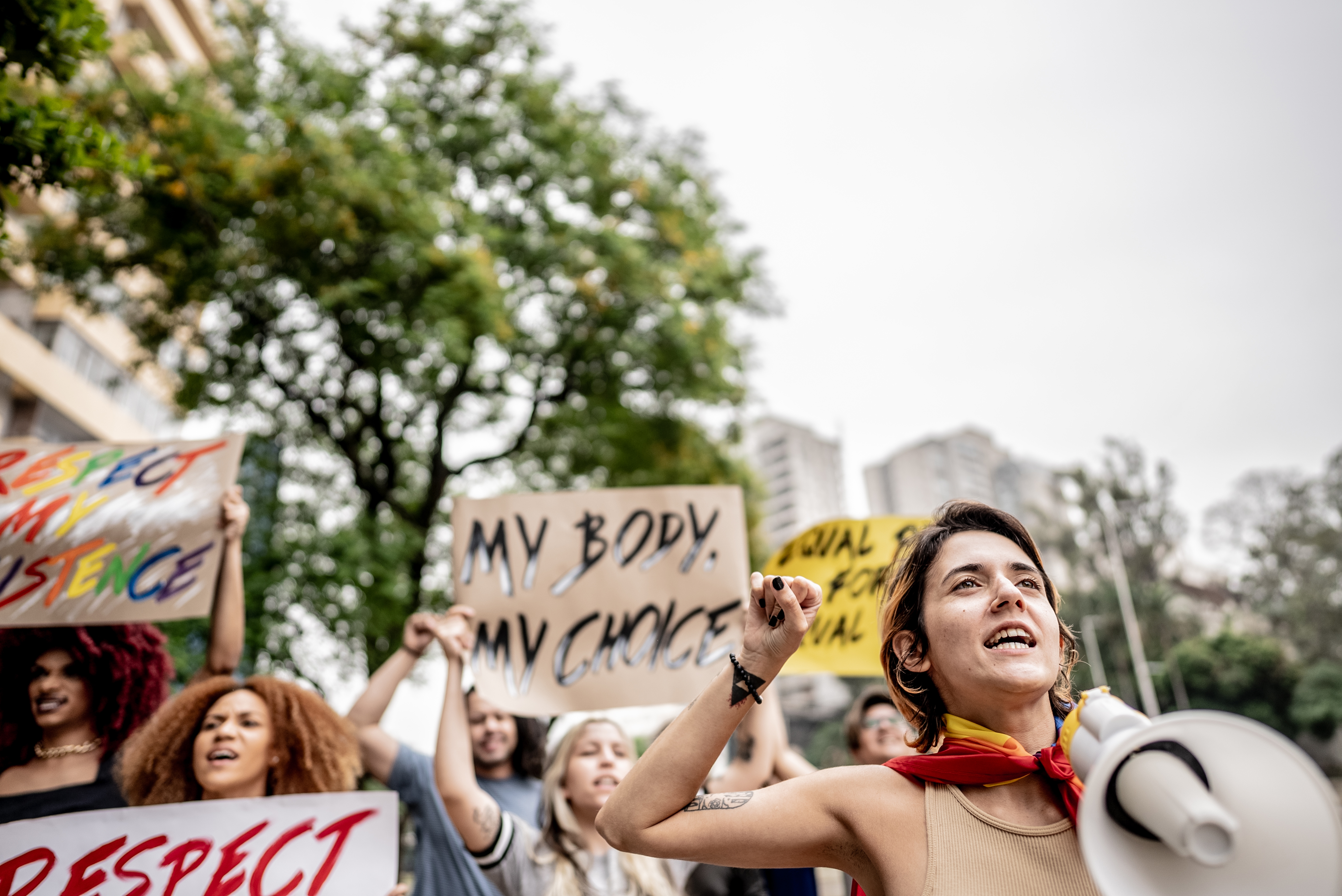 People at a protest holding signs, one reads "My Body, My Choice." A person in front has a megaphone, wearing casual attire with a scarf
