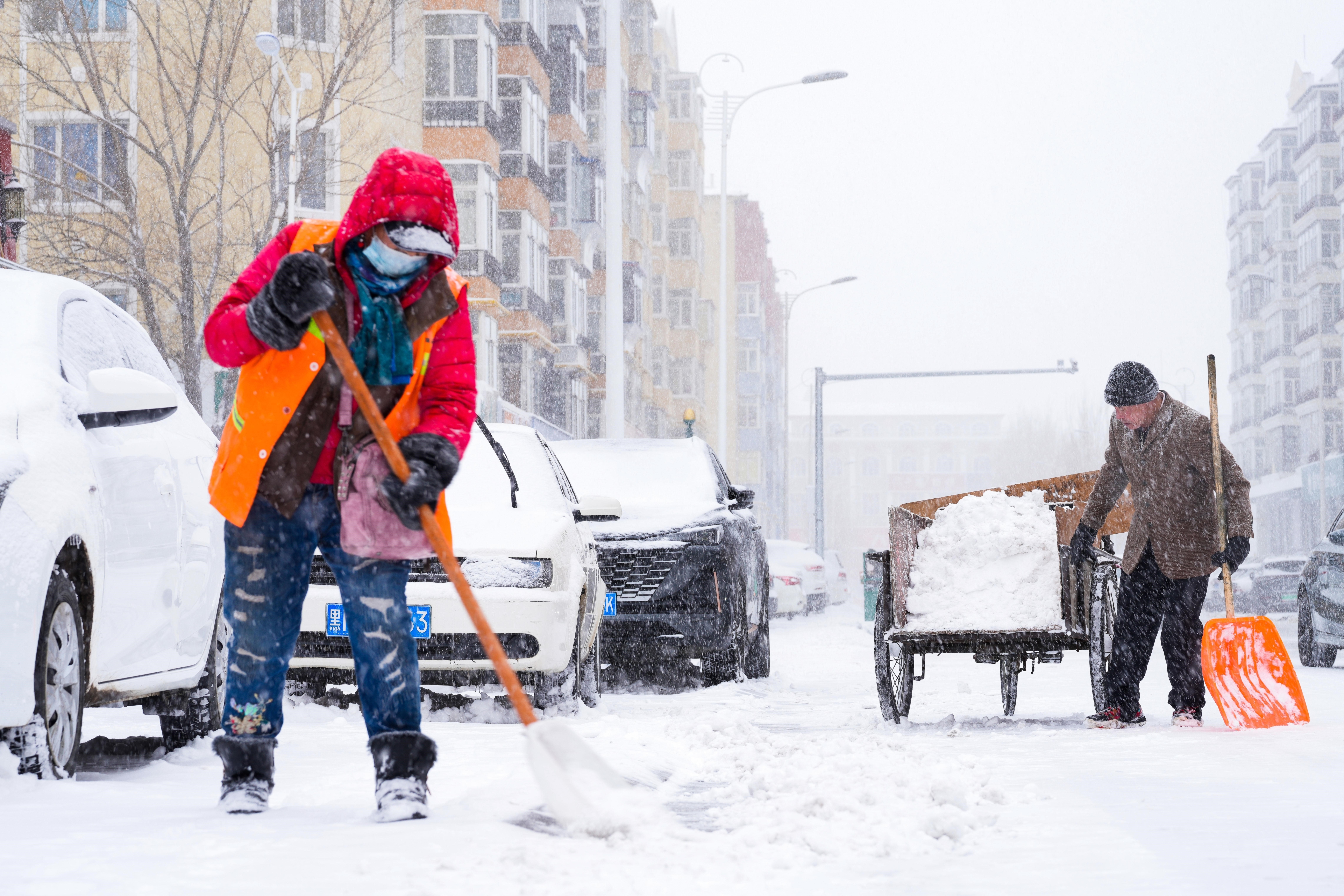 Two people shoveling snow on a city street during a snowstorm, with buildings and parked cars in the background