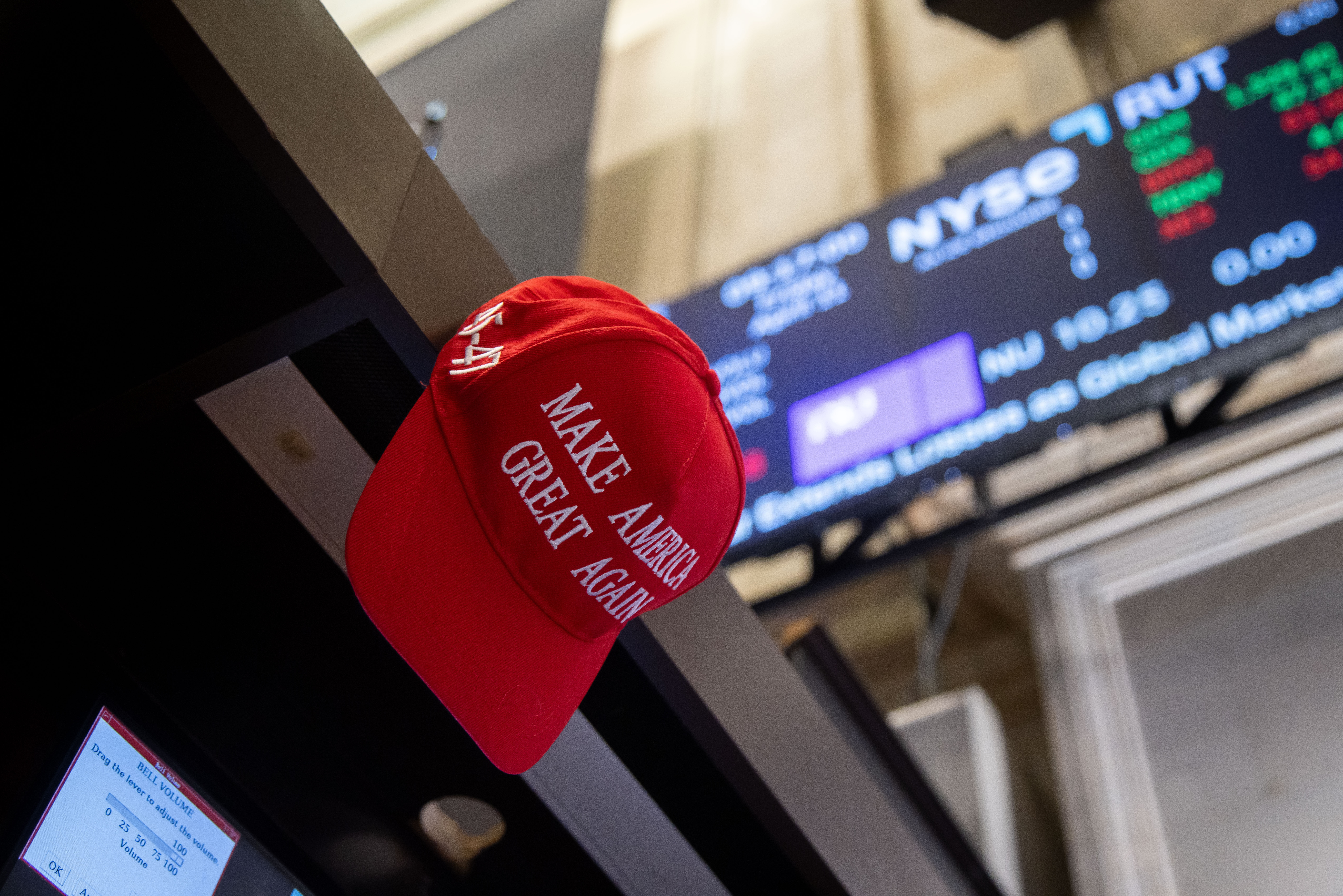 Red "Make America Great Again" hat on display inside a stock exchange, with electronic stock board showing numbers in the background