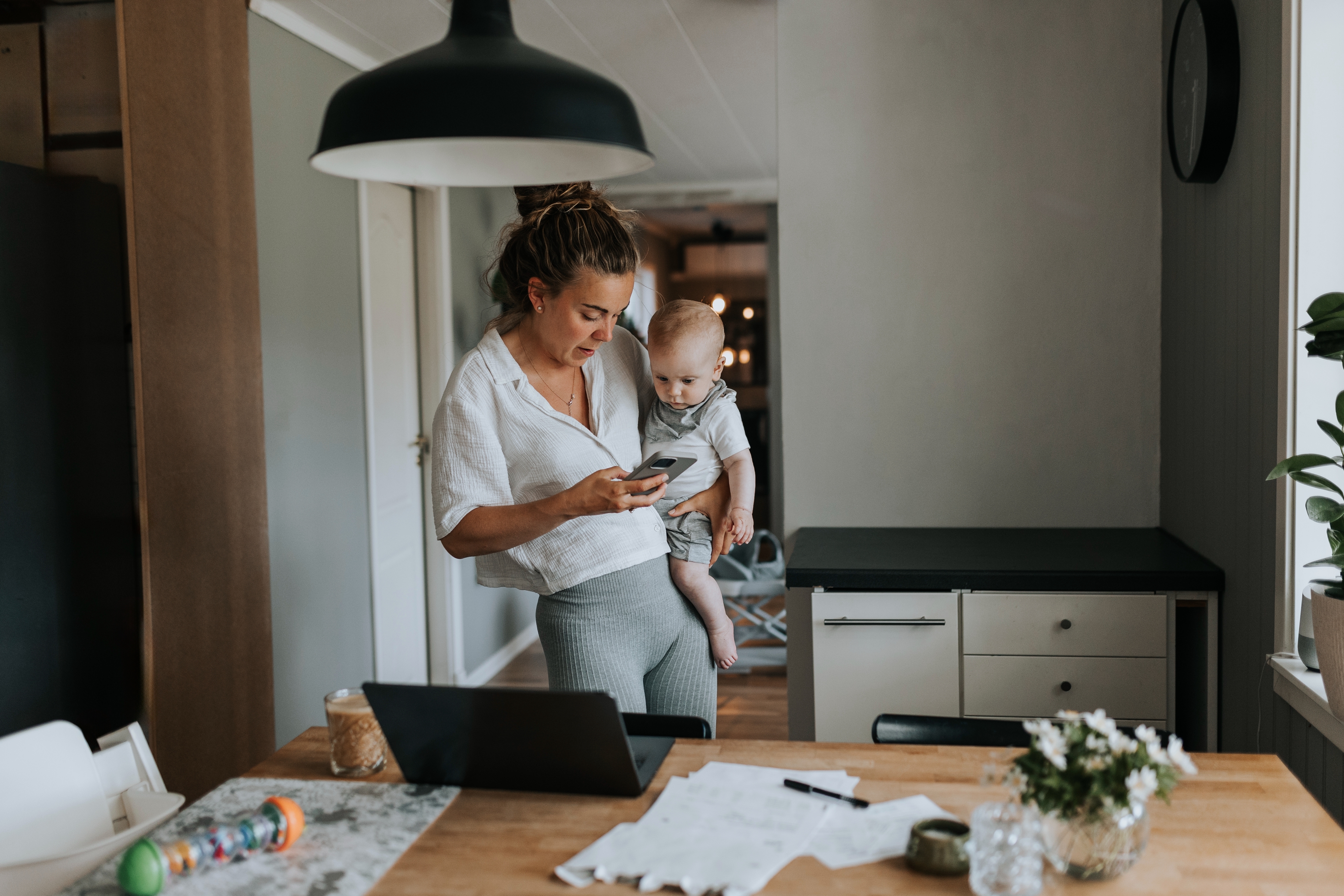 A woman holding a baby checks her phone while standing in a modern kitchen, with a laptop and documents on the table
