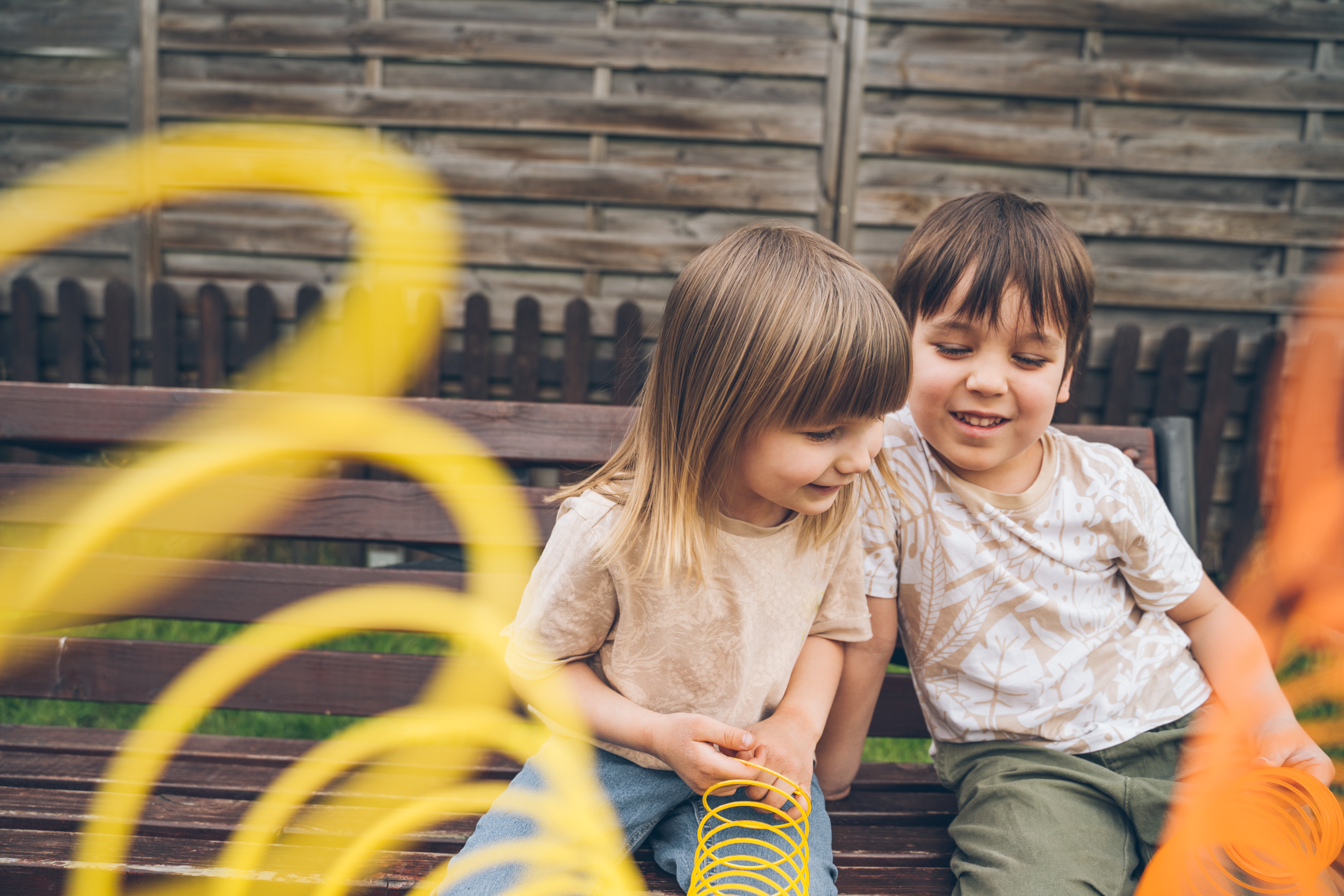 Two young children play with large coiled toys on a wooden bench in a garden. They both have joyful expressions
