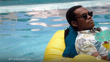 A person relaxes on a yellow pool float wearing sunglasses and a tropical shirt. Text in corner: #TheGoodPlace, NBC