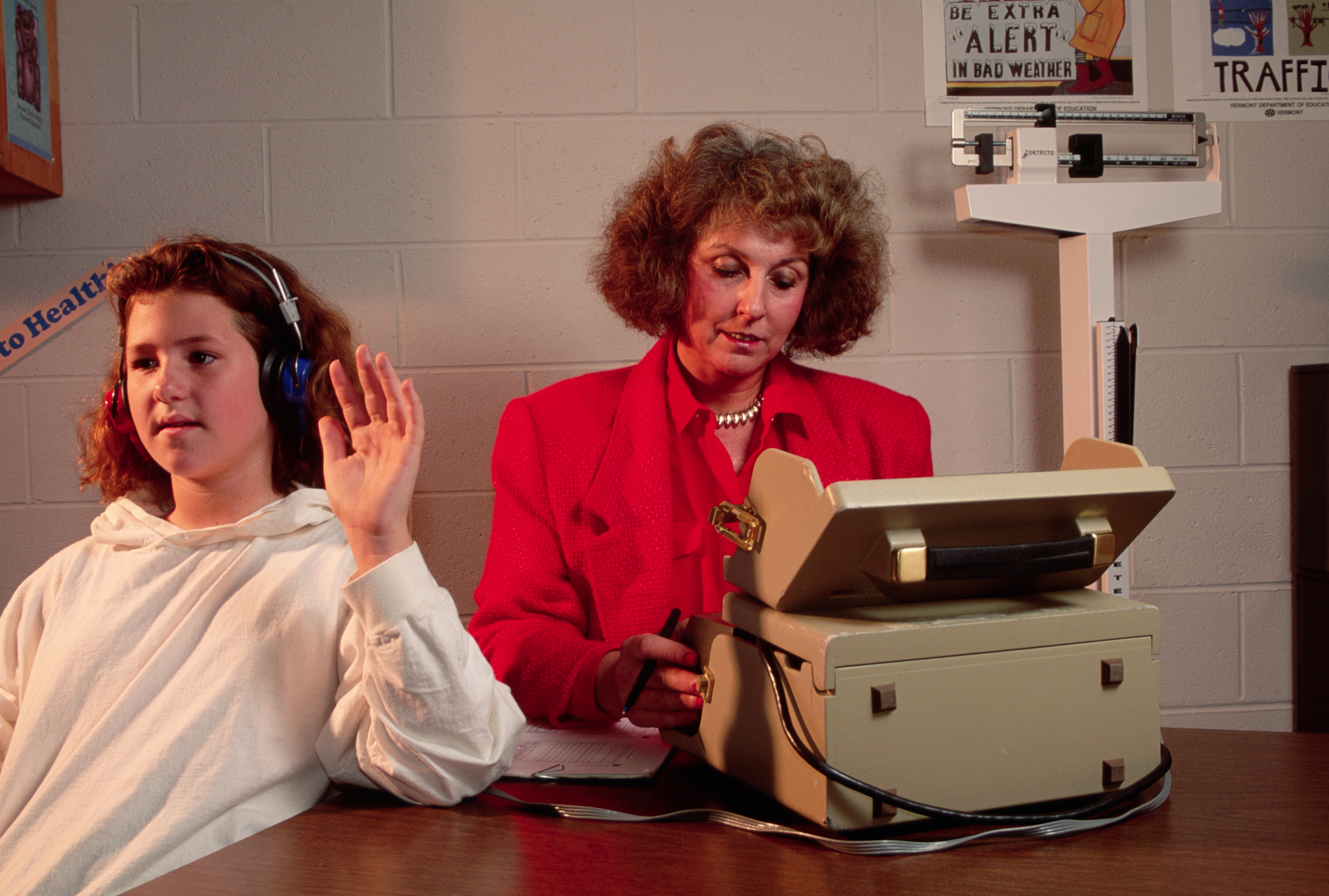 A child wearing headphones raises their hand during a hearing test, while an adult operates an audiometer and takes notes