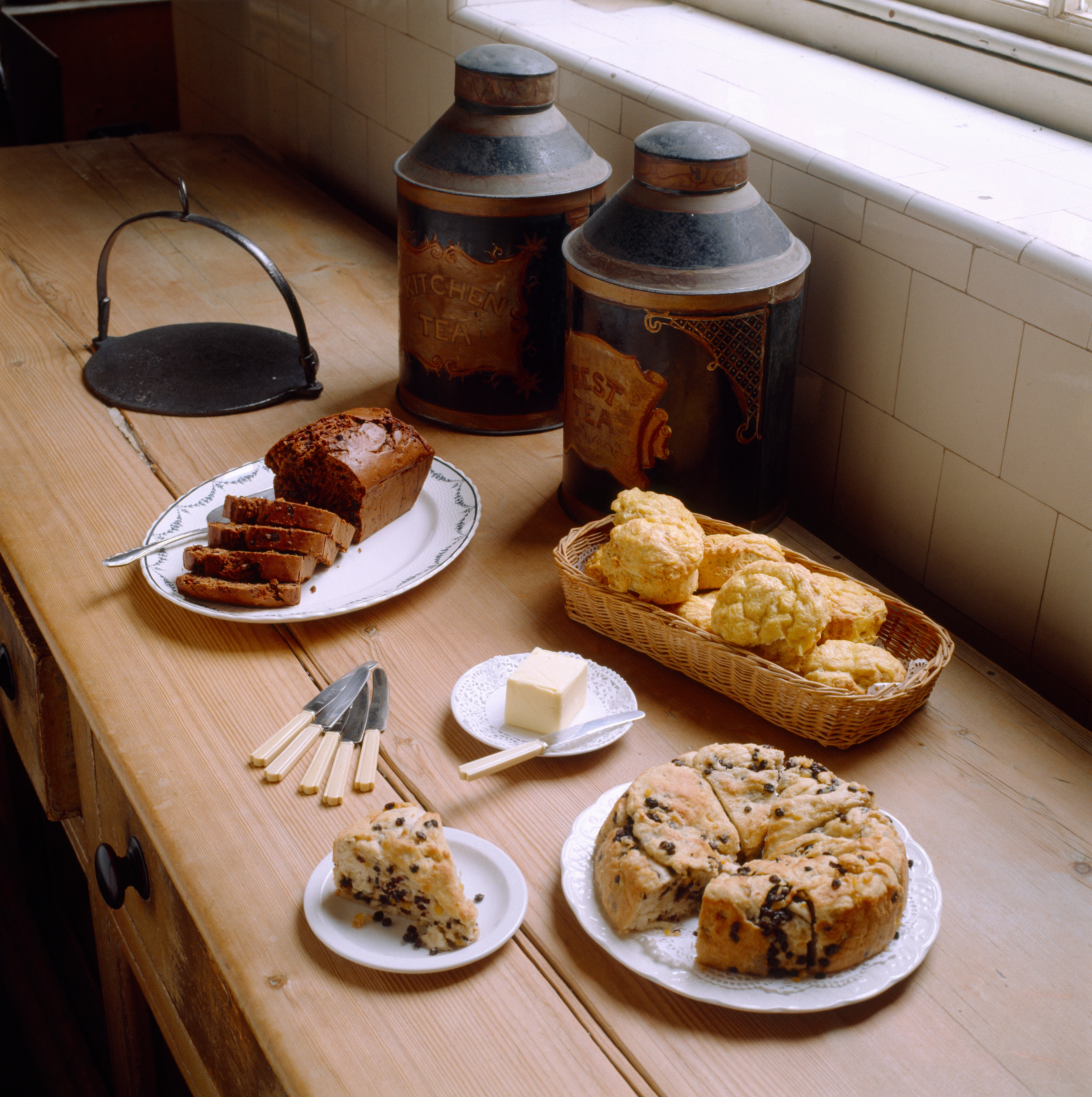 Assorted baked goods on a wooden counter, including sliced bread, scones, and pastries next to butter with vintage cannisters in the background