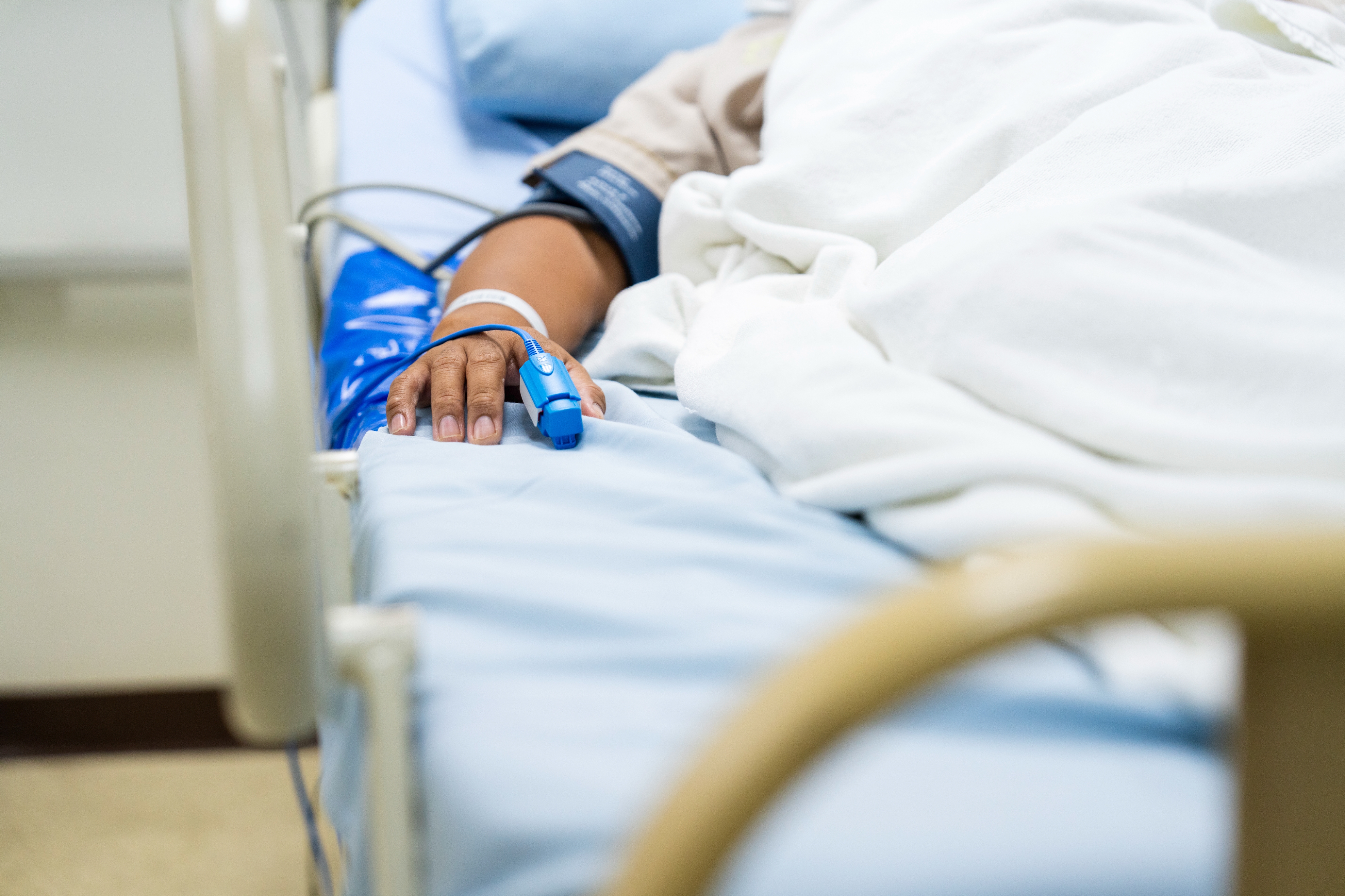 Person in hospital bed with medical monitor on finger, IV line, and blood pressure cuff on arm, symbolizing medical care and monitoring