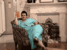 Person in a blue robe sits casually with a drink on a patterned chair, exuding a relaxed, confident demeanor