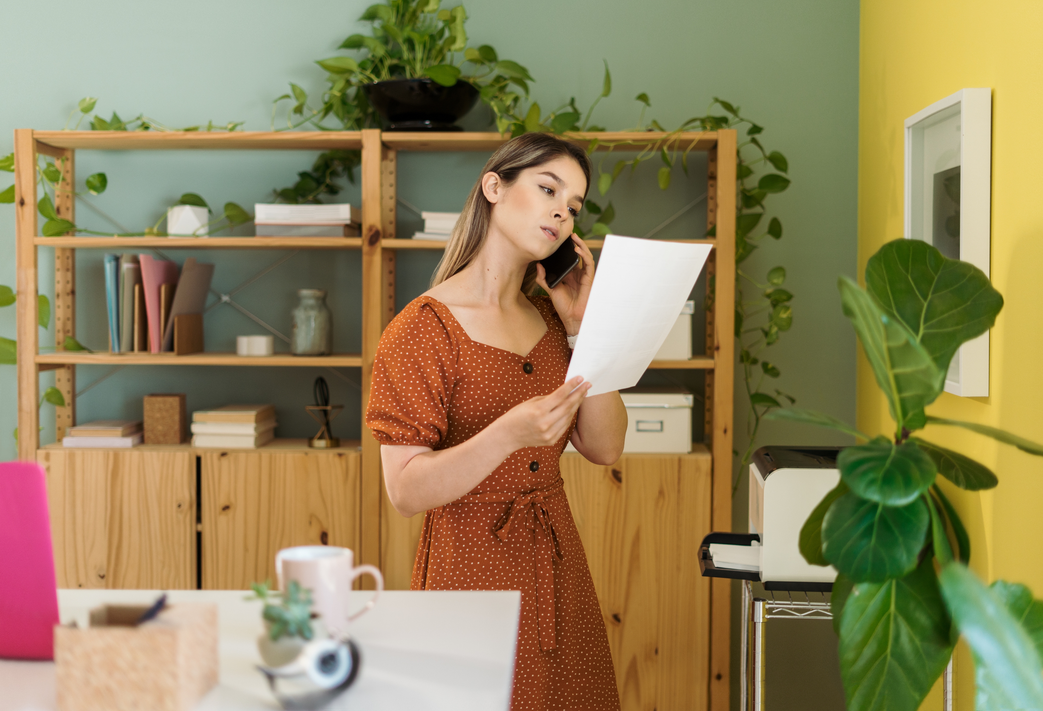 Person in polka dot dress reads a paper in an office with plants and bookshelves, creating a cozy, thoughtful atmosphere