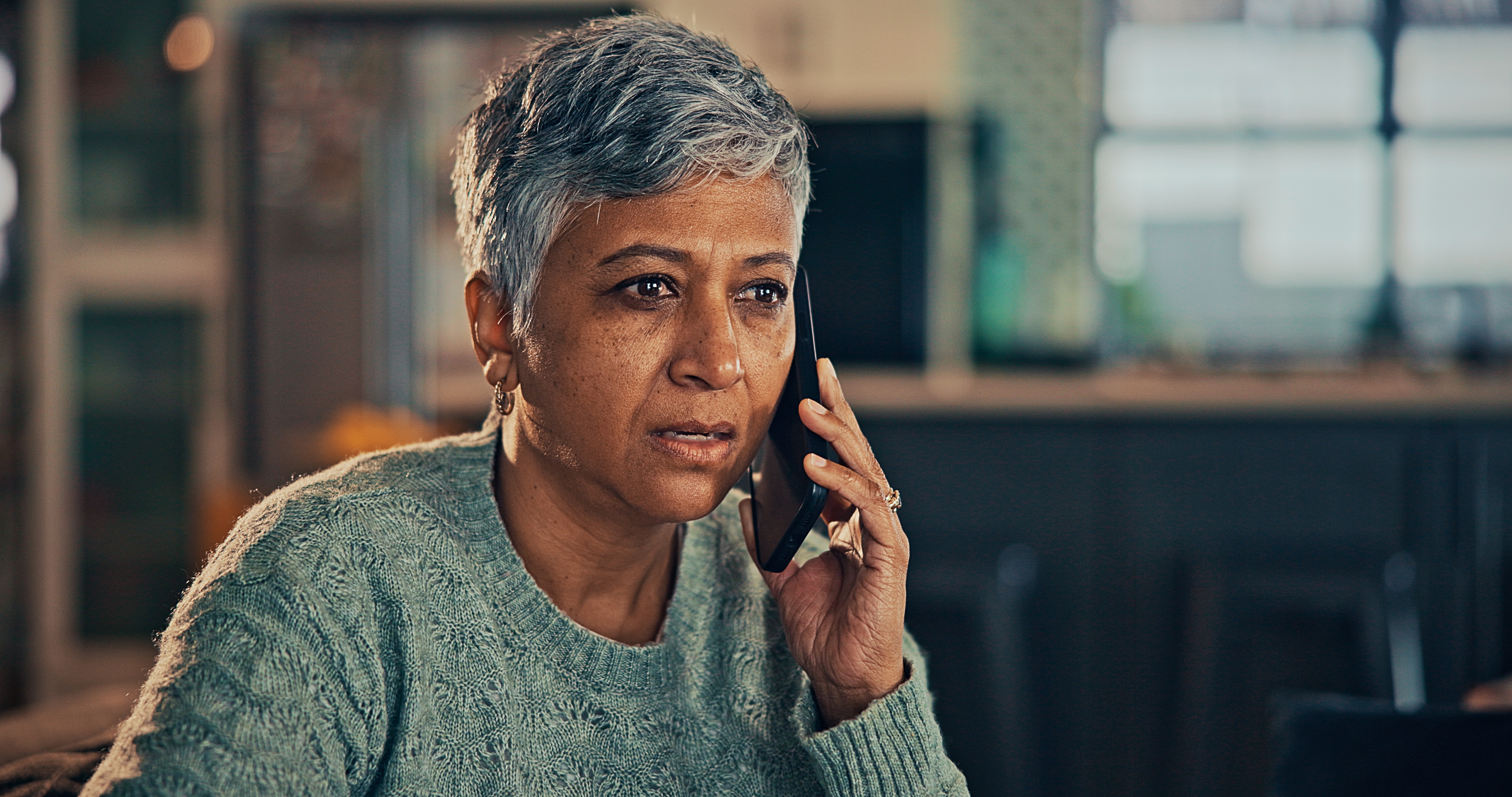 Person with short hair, wearing a textured sweater, talking on the phone with a concerned expression in an indoor setting