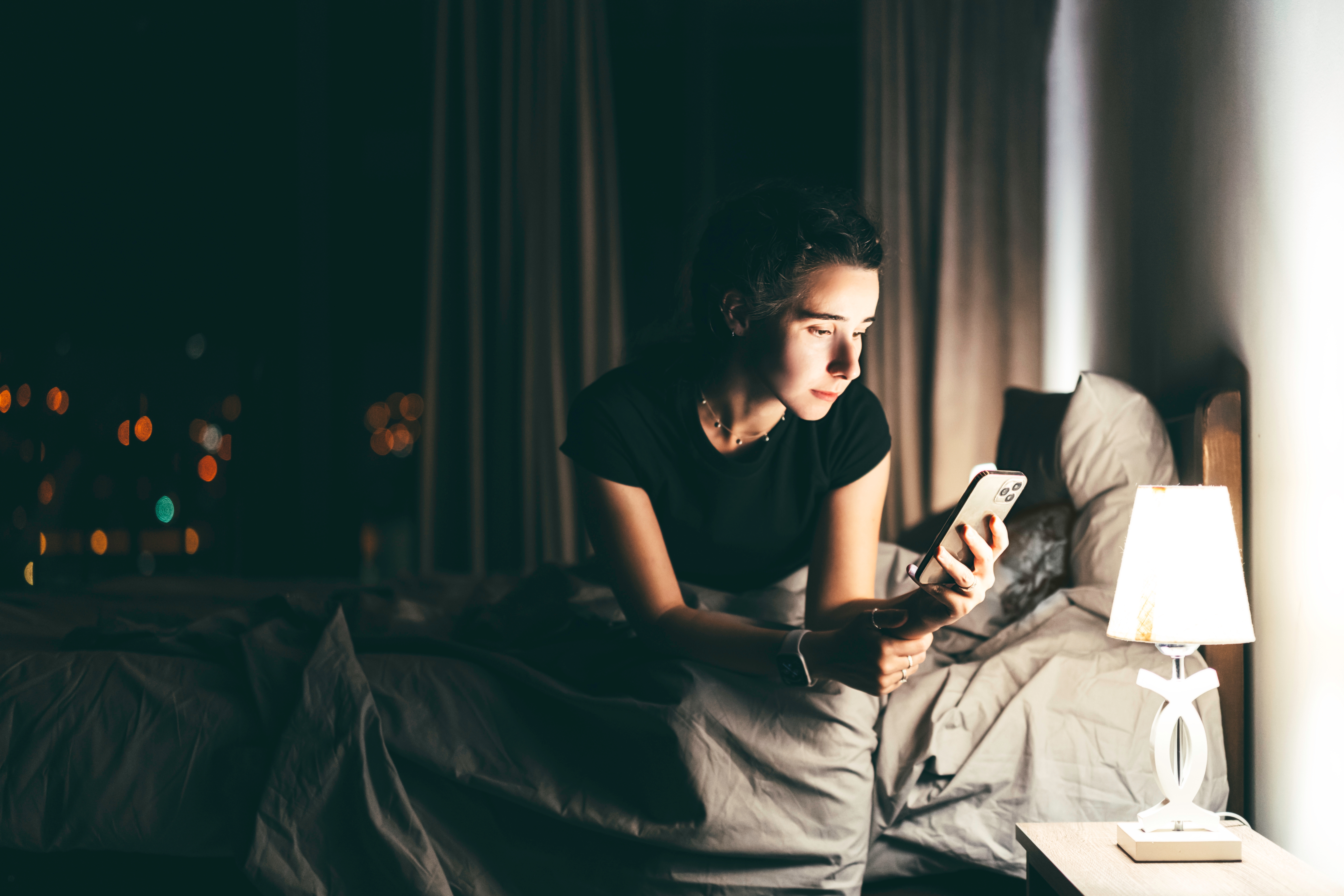 Person in bed at night looking at a smartphone, illuminated by a nearby lamp, suggesting late-night communication or reflection