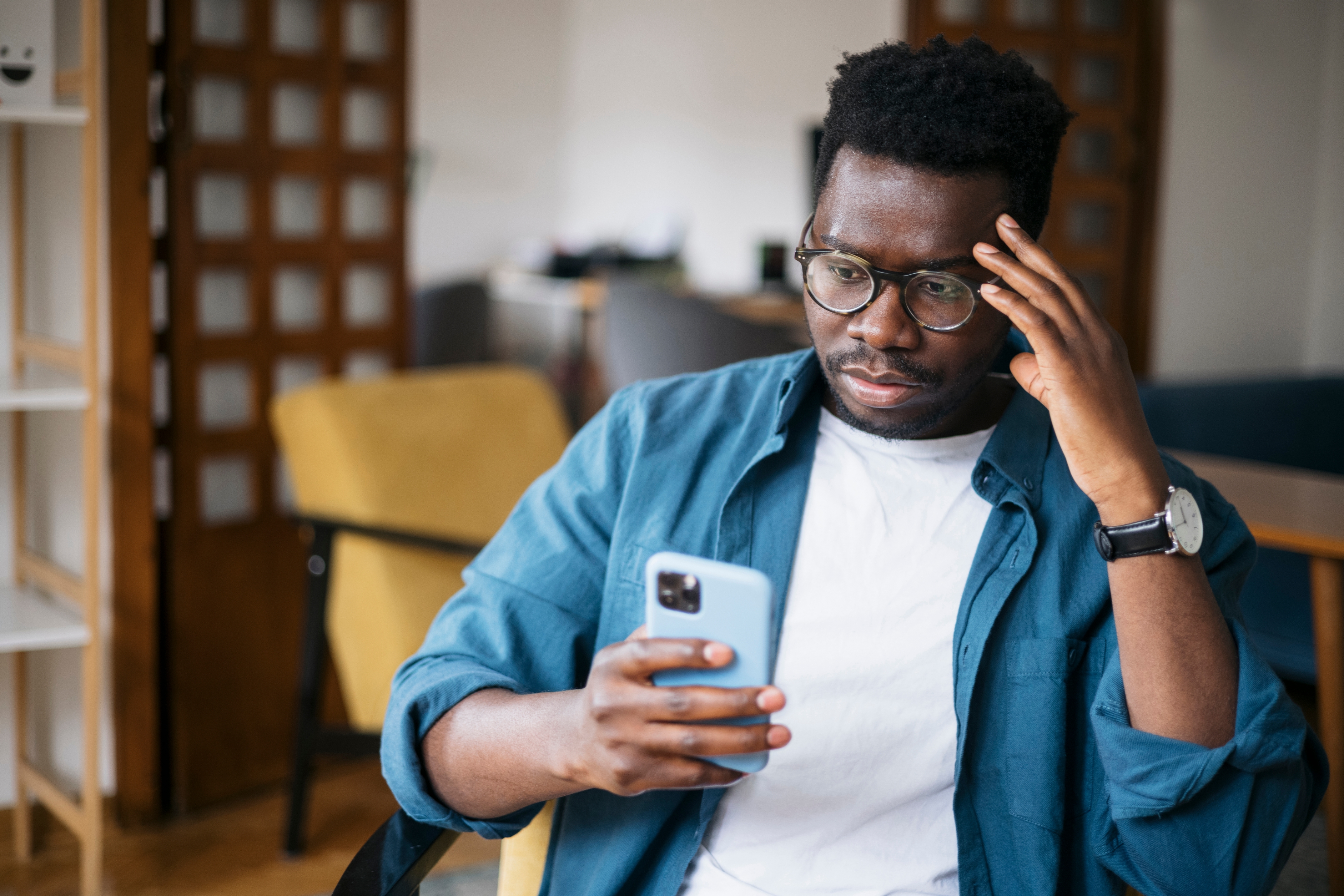 Person wearing glasses and a blue shirt, sitting and looking at a phone with a thoughtful expression, hand on temple