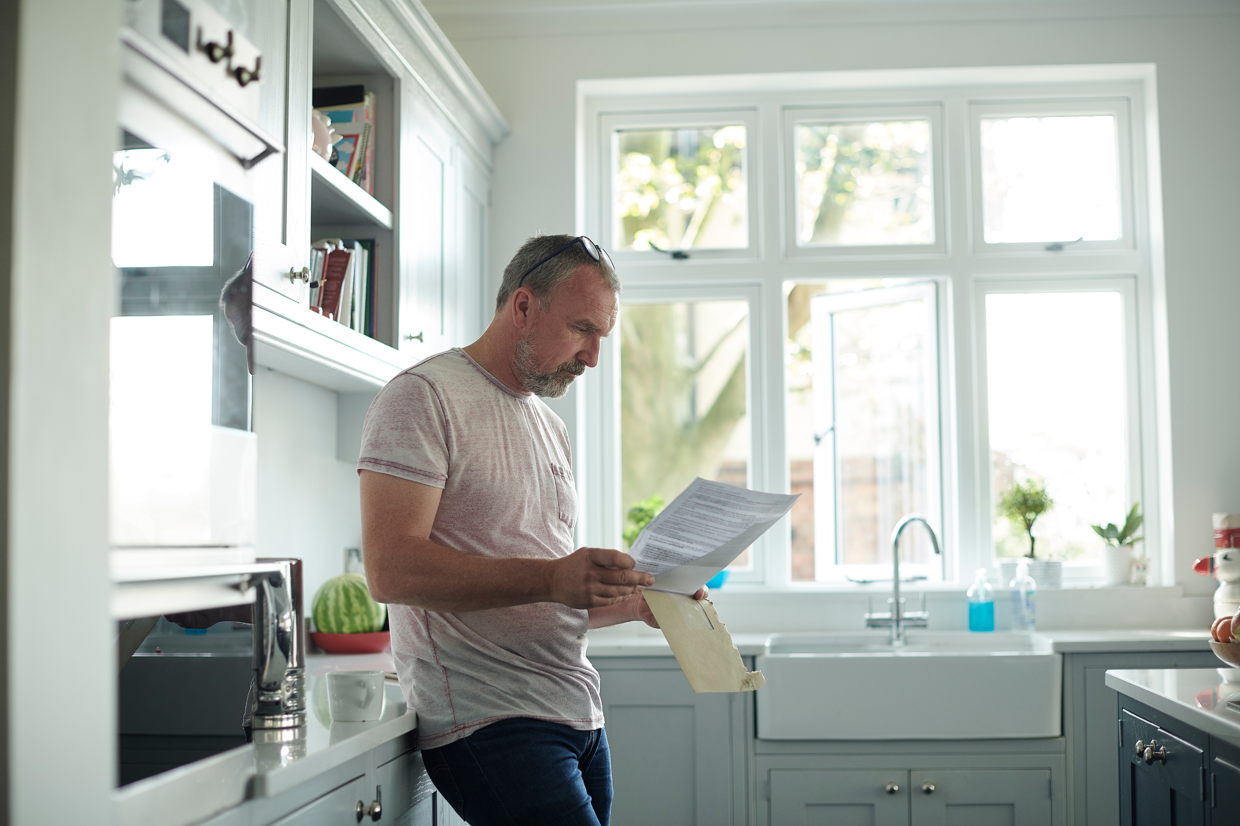 Man in kitchen thoughtfully reading a letter, leaning against a counter near a window with natural light streaming in